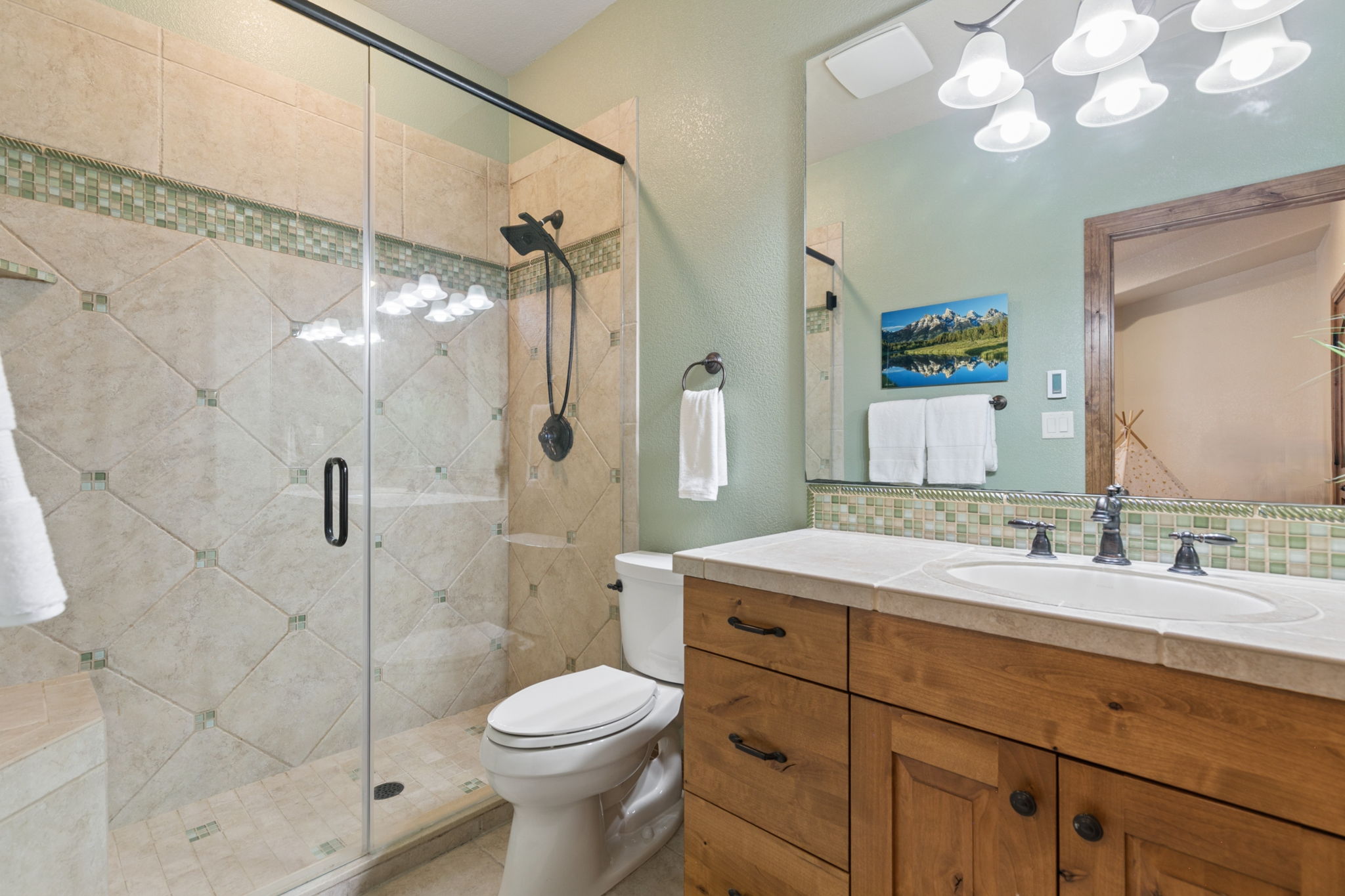 This bathroom features a walk-in shower with a glass door, tiled walls with decorative accents, and a light wood vanity with a stone countertop. A large mirror above the vanity reflects the room, including a piece of mountain landscape art on the sage green wall. The space is well-lit by a multi-bulb fixture and includes a standard toilet and towel storage.