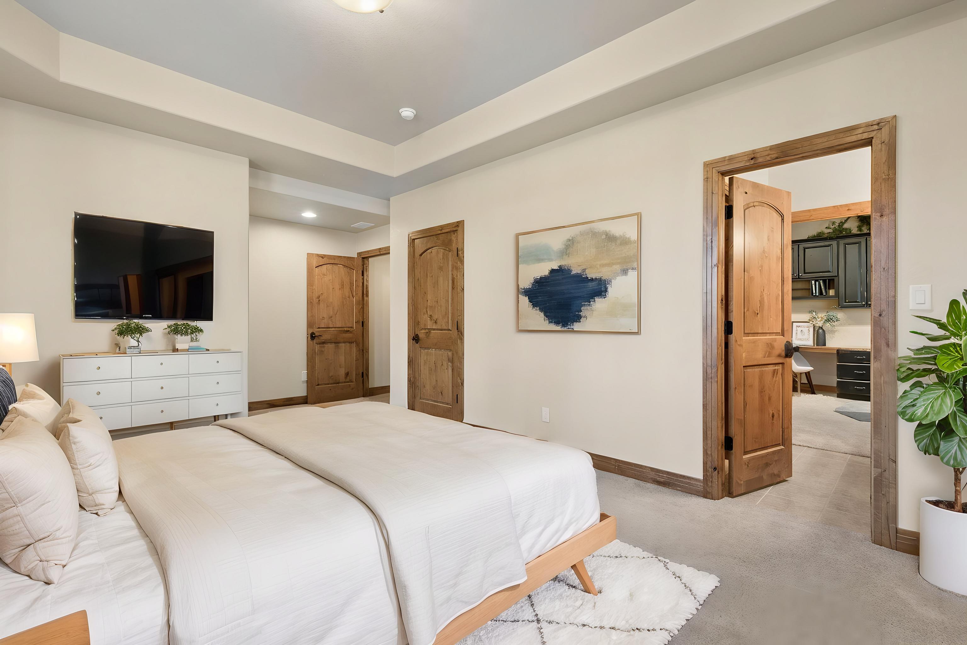 This primary bedroom features a neutral color palette with a light wood bed frame and a white dresser topped with a wall-mounted television. The room includes a tray ceiling, a piece of abstract wall art, and a doorway leading into an adjacent office space. A large potted plant adds a touch of greenery to the corner, while the overall aesthetic is clean, bright, and inviting.
