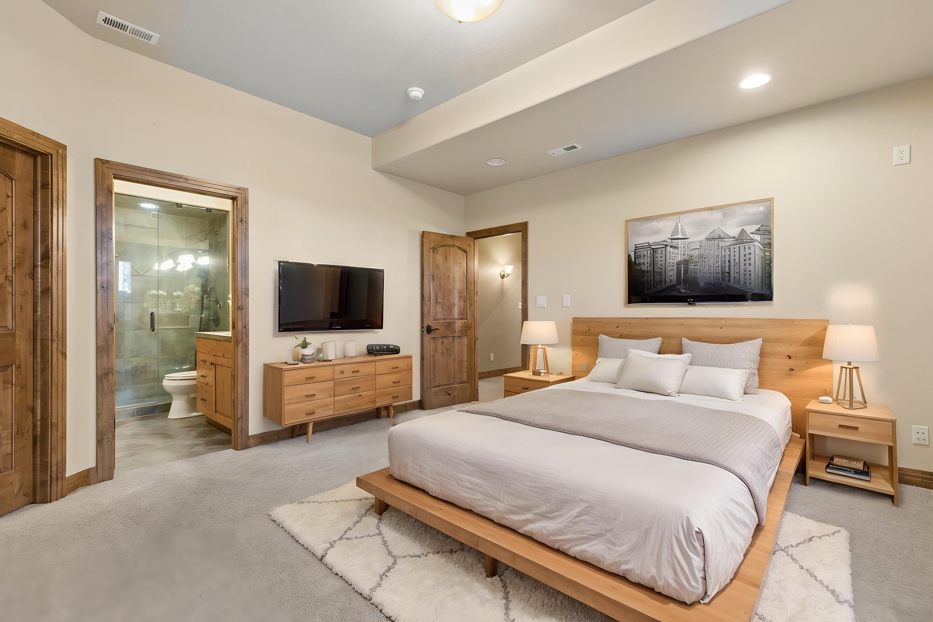 This primary bedroom features a platform bed with a light wood headboard, complemented by matching nightstands and a dresser. The room is decorated with a large black-and-white cityscape print above the bed and a textured area rug over neutral carpeting. A doorway to the left provides a view into an attached bathroom, while the overall aesthetic is clean, modern, and inviting.