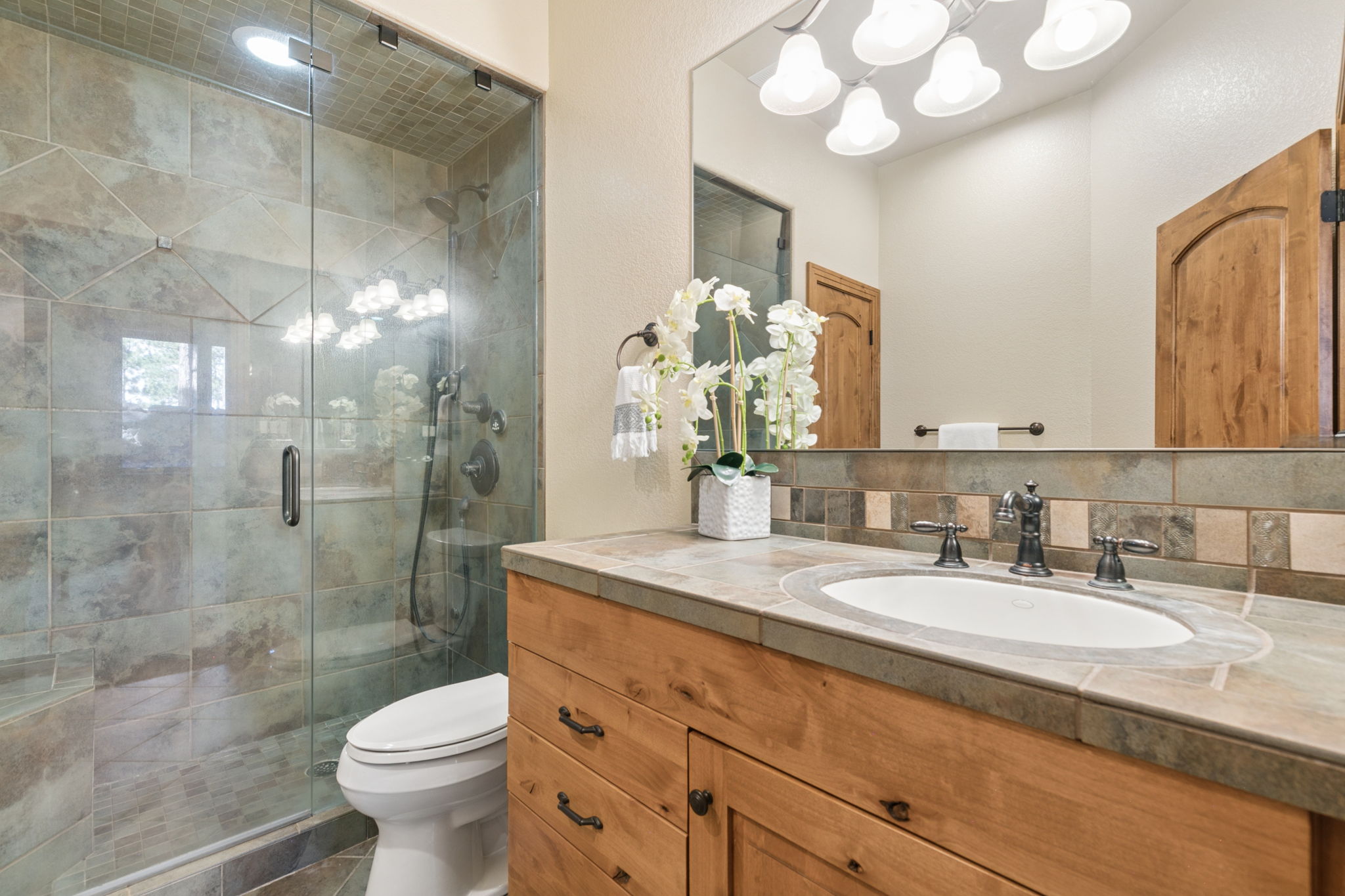 This bathroom features a rustic aesthetic with a light wood vanity topped with a stone counter and a white undermount sink. To the left, a glass-enclosed shower is finished with textured, earth-toned tiles, while a large mirror above the vanity reflects the room's warm lighting. A white toilet is positioned beside the vanity, and a decorative floral arrangement adds a touch of elegance to the space.