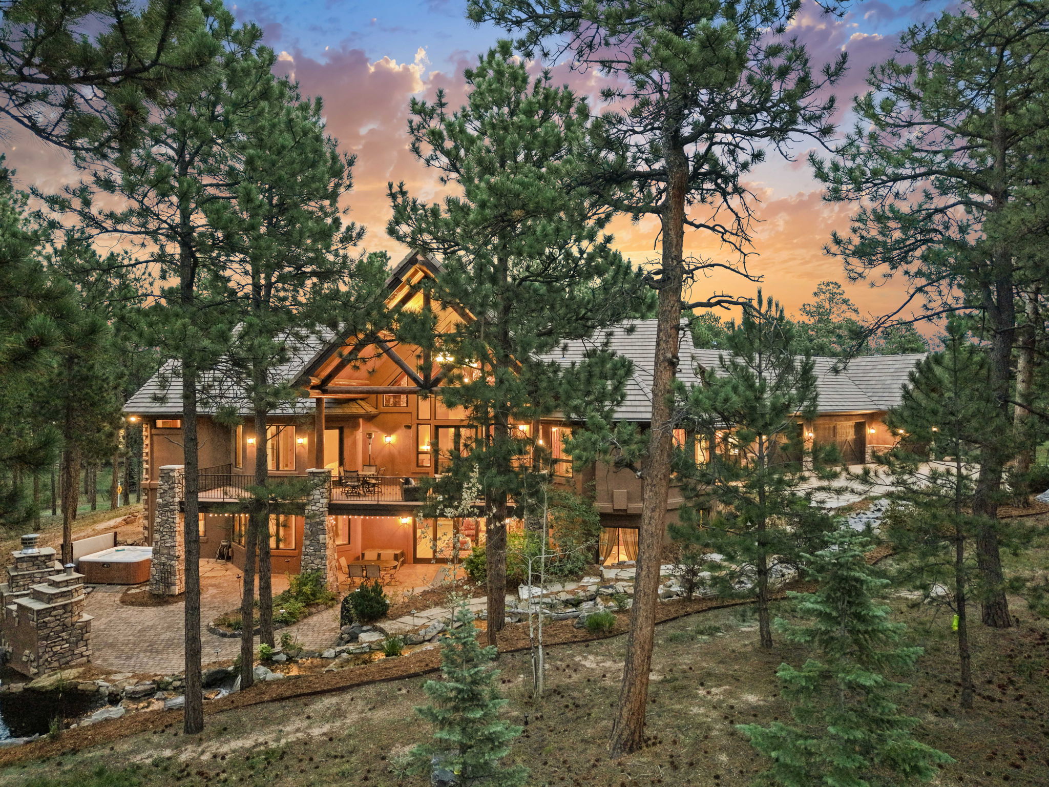 This expansive mountain-style residence is captured from an elevated rear perspective at dusk, showcasing a multi-level design with warm exterior lighting. The home features a prominent timber-framed gable, a spacious upper deck, and a lower-level stone patio complete with a hot tub and landscaped rock features. Surrounded by mature pine trees, the property exudes a serene, secluded atmosphere perfect for luxury mountain living.