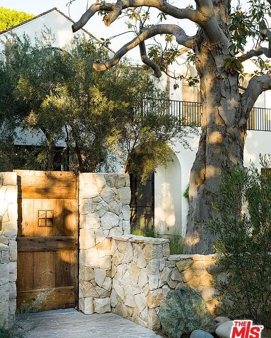 This image showcases an enchanting entryway, featuring a rustic wooden gate set within a stone wall. Mature trees flank the entrance, adding to the serene and private atmosphere. The combination of natural materials and lush greenery creates a charming and inviting curb appeal.