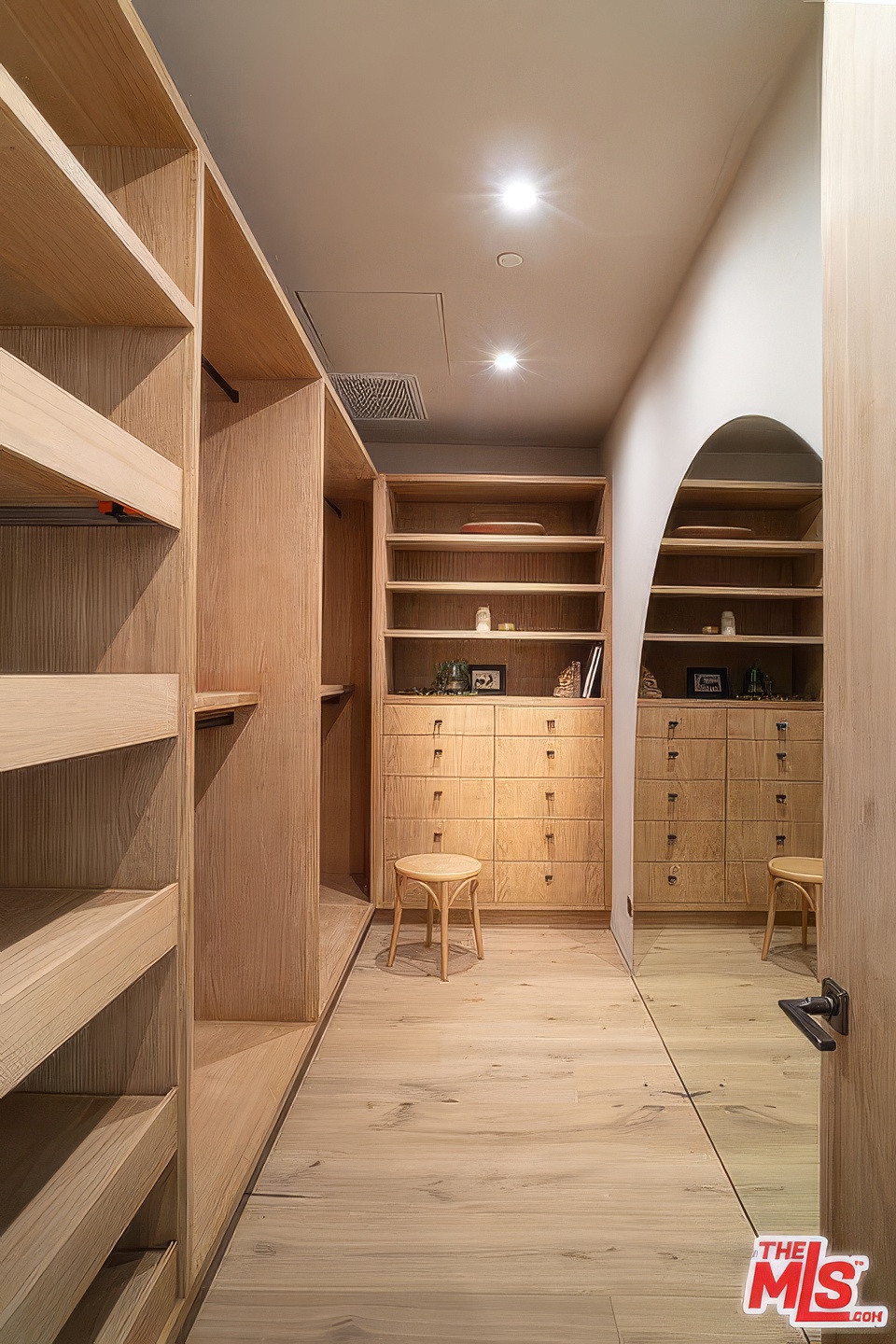 This is an interior shot of a well-organized walk-in closet. The closet features natural wood shelving and drawers, providing ample storage space. A stool is present, and a mirror with an arched top adds visual interest and functionality to the space.