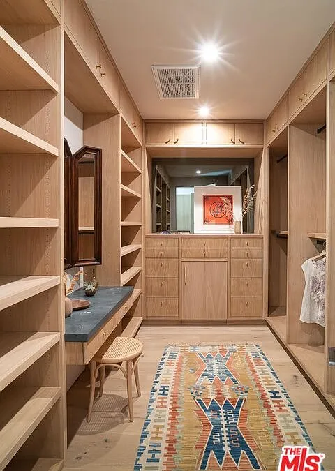 This image showcases a well-organized walk-in closet. It features custom-built wooden shelving and drawers, a vanity section with a stool, and a patterned area rug. The closet appears spacious and designed for optimal storage and organization.
