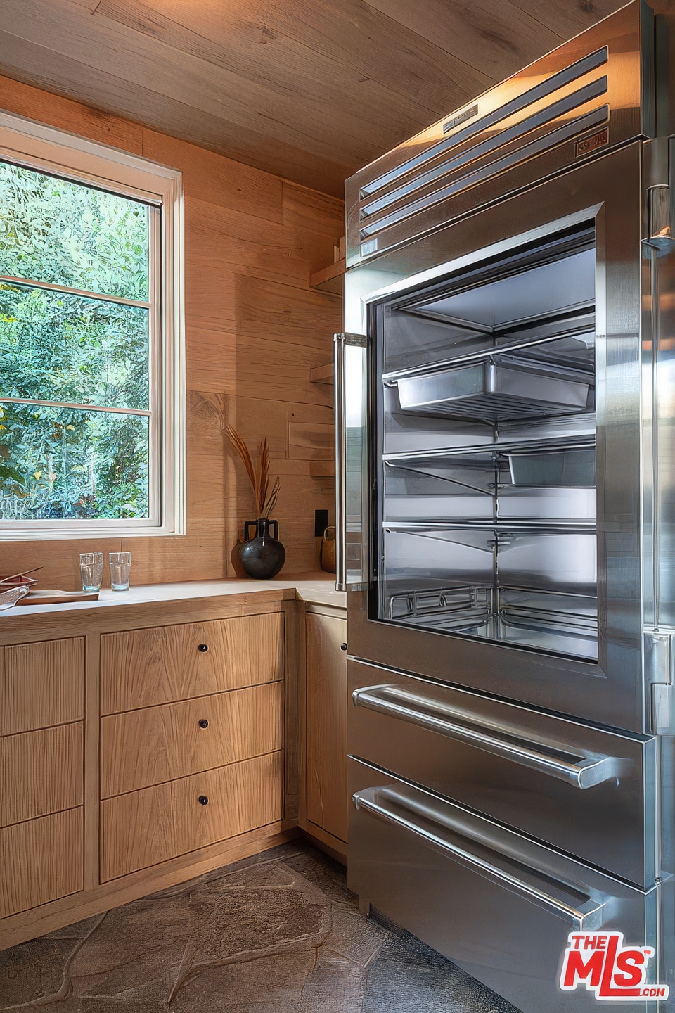 An interior shot of a kitchen features light wood cabinetry and a stainless steel refrigerator with a glass door. The floor is made of stone tiles, and a window allows natural light to enter. The kitchen's design incorporates natural elements and modern appliances.