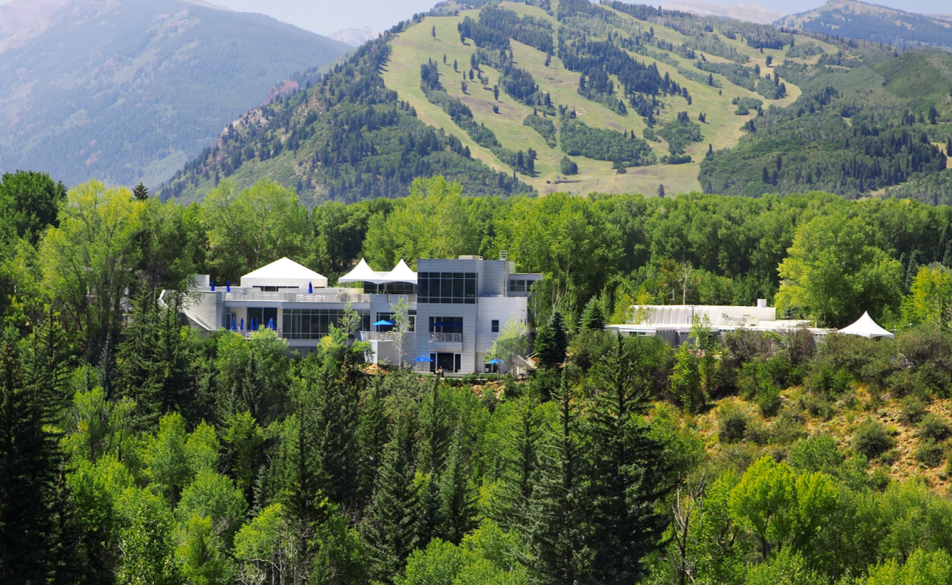 This image features a modern, multi-level building complex nestled into a lush, forested landscape with sweeping mountain views. The architecture combines contemporary glass and metallic elements with geometric white tent structures, suggesting a high-end mountain resort, event center, or community clubhouse. The setting offers a serene, elevated atmosphere, blending luxury amenities with the natural beauty of the surrounding wooded hillside.