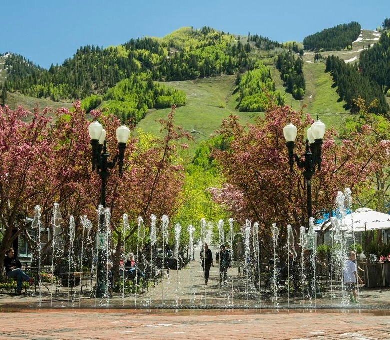 This picturesque outdoor community area features a vibrant public plaza adorned with blooming cherry blossom trees and an interactive water fountain feature. The setting captures a beautiful mountain backdrop, providing an inviting and upscale atmosphere characteristic of a resort-style neighborhood or plaza. It is a highly desirable space for residents and visitors to enjoy leisure time amidst scenic natural surroundings.