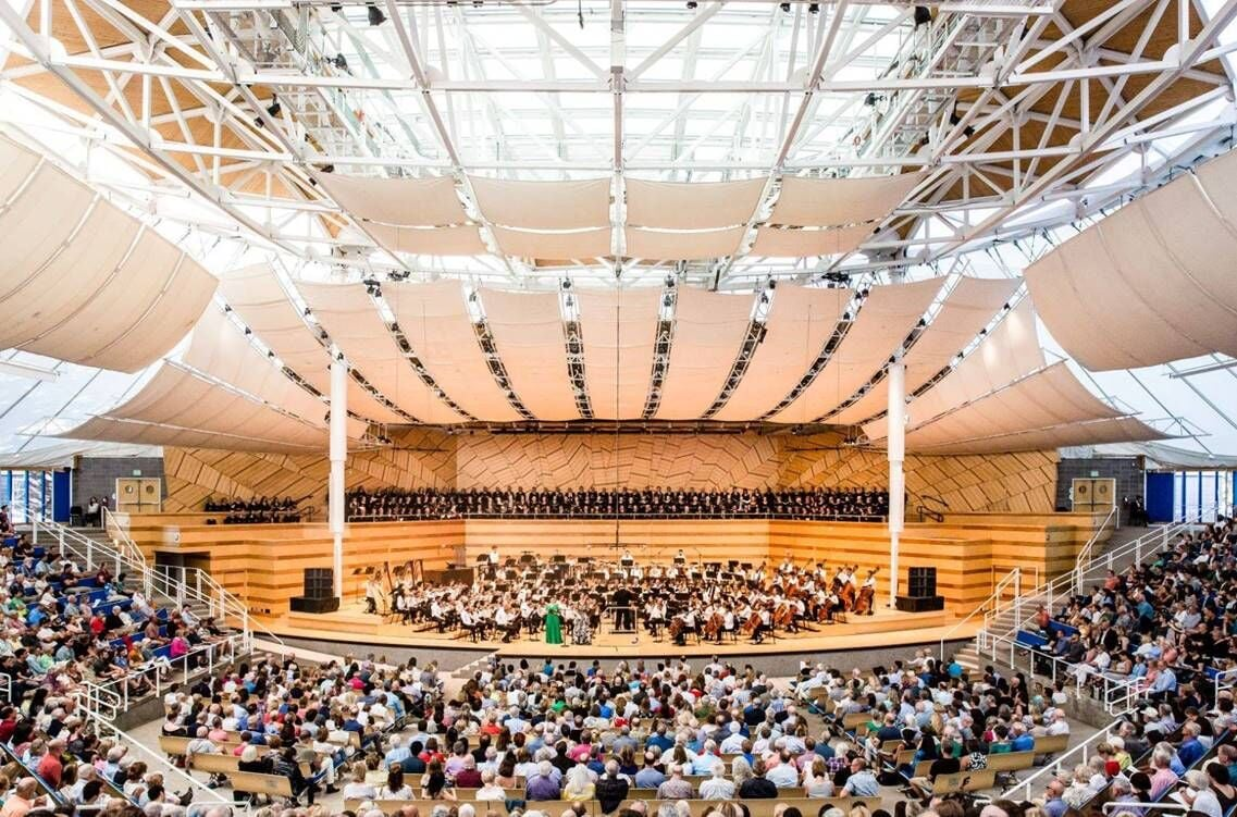 This image depicts a large, open-air concert venue or amphitheater characterized by its striking modern architectural roof design composed of steel trusses and draped fabric panels. The space features tiered seating for a large audience, a broad performance stage for an orchestra and choir, and warm wood-paneled walls, creating an inviting and professional atmosphere for public performances.