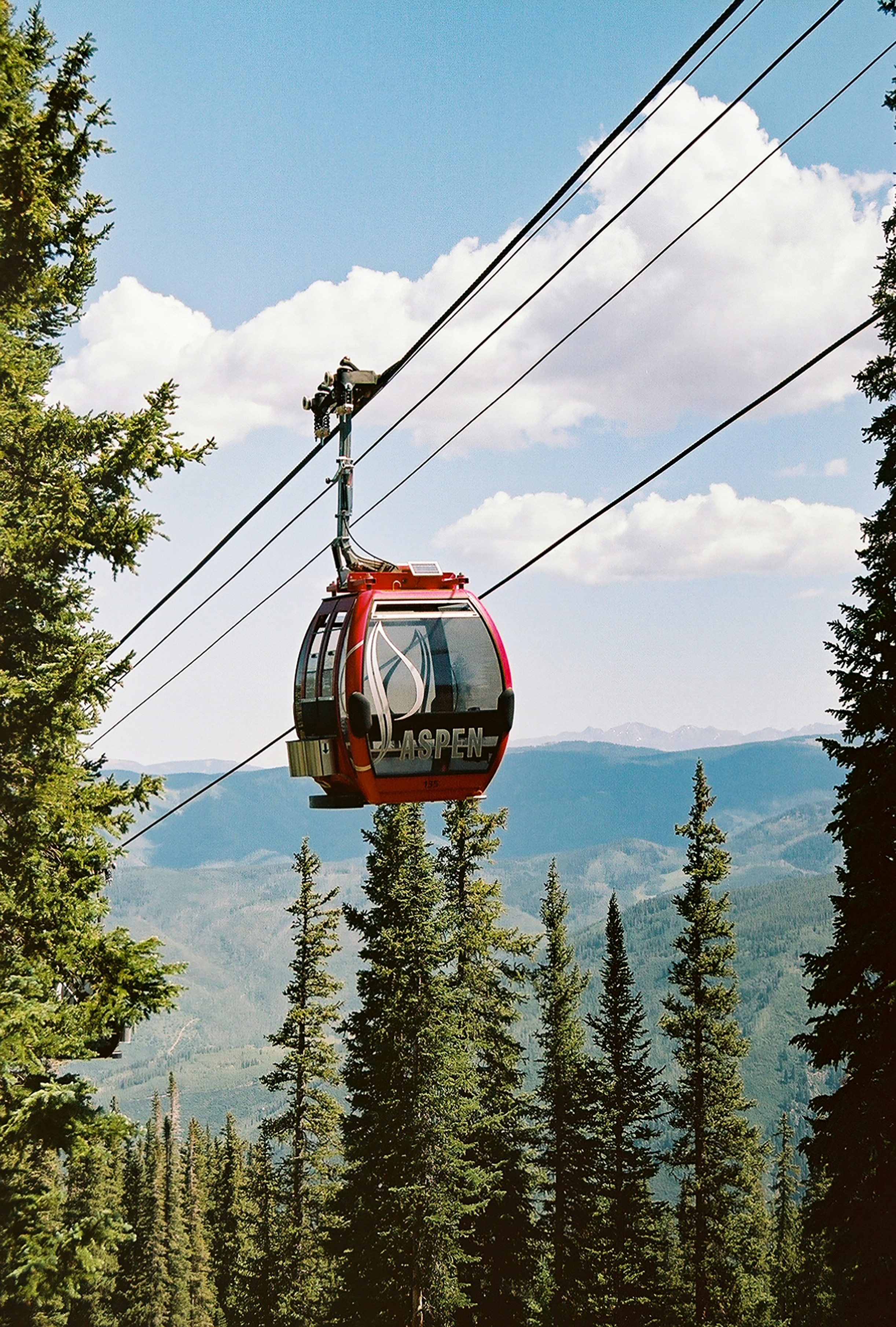 This image features a red gondola car, marked with 'Aspen', operating on a cable system against a stunning mountainous backdrop. It highlights the premium recreational amenities associated with living in or visiting a major mountain resort destination. The scene provides a direct connection to high-end outdoor leisure activities and scenic mountain views highly valued in luxury real estate.