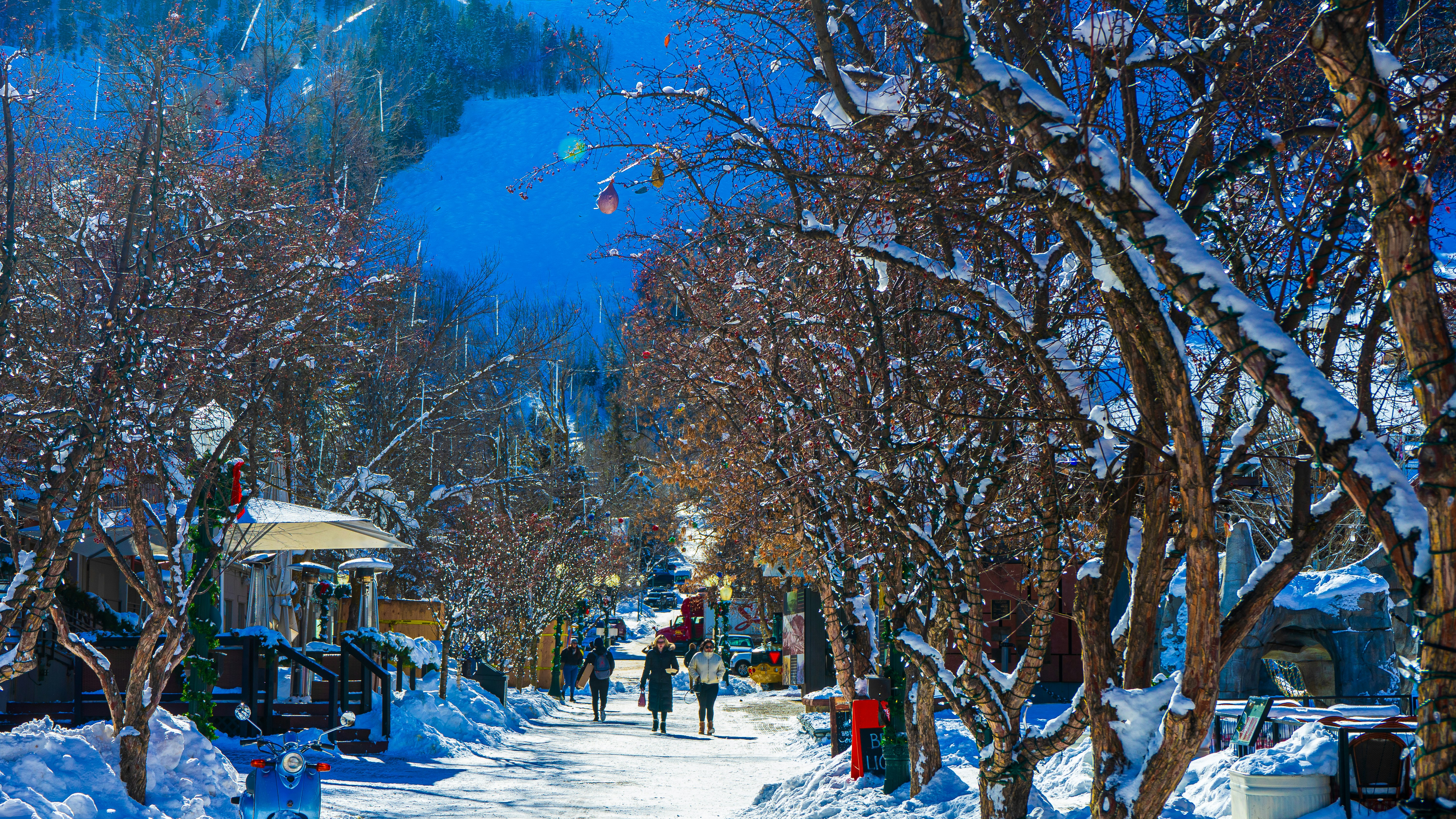 This winter scene depicts a charming, walkable commercial corridor in a snowy alpine resort setting, characterized by tree-lined paths and adjacent outdoor dining areas. The street is blanketed in fresh snow, flanked by rustic buildings and mature trees decorated with string lights, creating an inviting village atmosphere. This location offers excellent pedestrian access to local amenities, perfect for a high-end ski destination lifestyle.