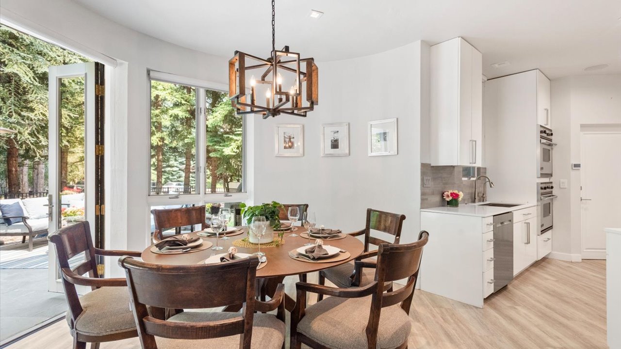 This bright dining area features a round wooden table surrounded by six upholstered chairs, positioned beneath a distinctive geometric chandelier. The space flows seamlessly into a modern kitchen with white cabinetry, stainless steel appliances, and light wood-toned flooring. Large windows and a glass door provide ample natural light and a view of the lush greenery outside, creating an inviting and open atmosphere.