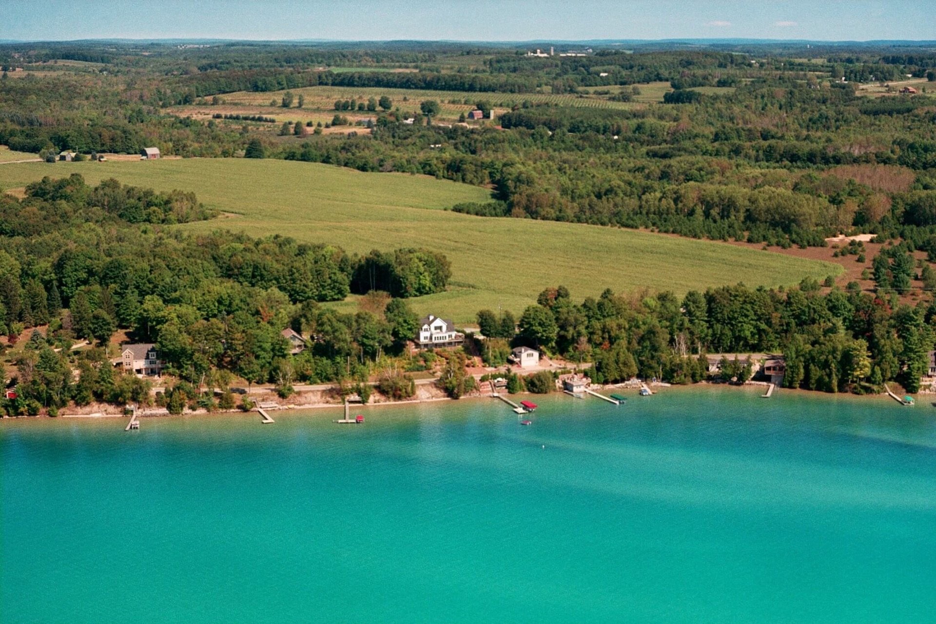 This aerial shot showcases a beautiful waterfront property with several homes lining the edge of a vibrant turquoise lake. The properties feature docks and waterfront access, nestled among lush greenery and trees. The landscape extends into the distance with rolling fields and forests, creating a serene and picturesque setting.