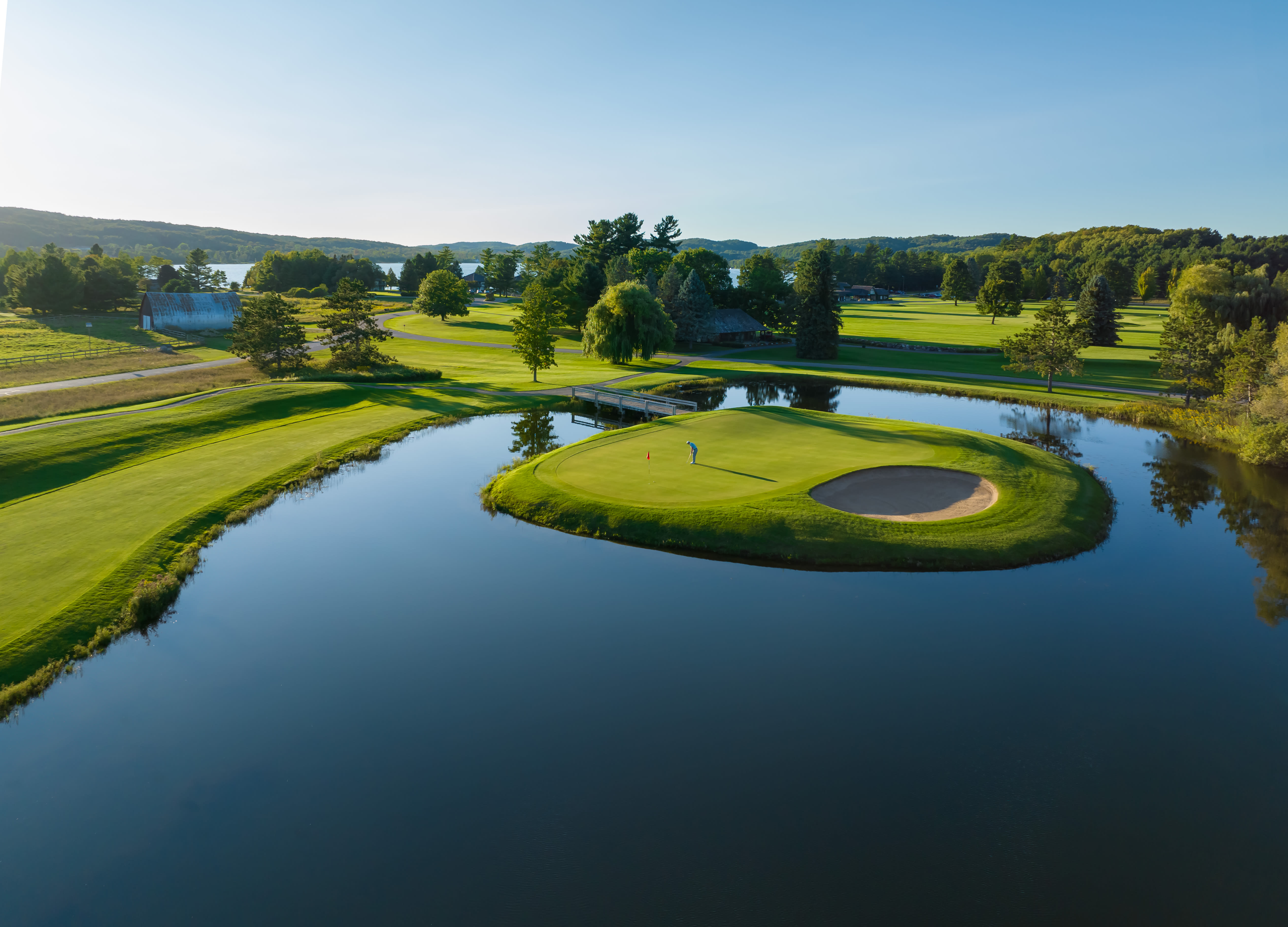This aerial view showcases a picturesque golf course featuring a green island surrounded by a tranquil pond. A golfer is visible on the island green, adding a sense of activity and scale. The surrounding landscape includes lush green fairways, trees, and water features, creating an inviting and scenic setting for potential buyers, indicative of a high-end community.