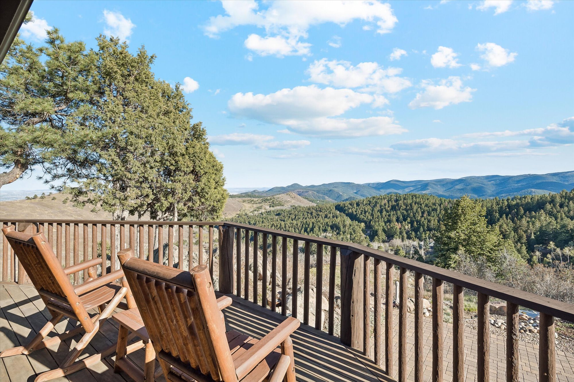 This elevated wooden deck features rustic log-style rocking chairs that provide a perfect vantage point for the scenic mountain landscape. A substantial wooden railing surrounds the perimeter, which overlooks a vast, forested valley under a bright, partly cloudy sky. The space offers a serene, panoramic outdoor living environment ideal for relaxation.