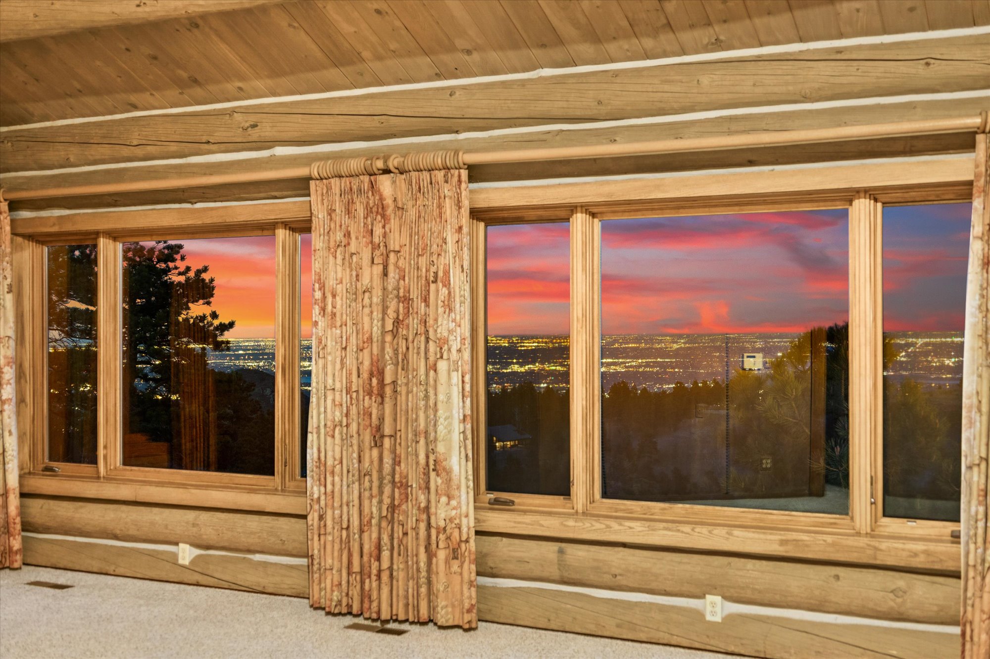 This living room space features rustic log cabin walls and a large, multi-pane window configuration that showcases a stunning, expansive panoramic sunset view over a distant city skyline. Neutral carpet flooring and patterned curtains complement the natural wood interior, creating a warm and inviting atmosphere. The impressive scenic vista is the focal point of the room, making it a standout feature for potential buyers.