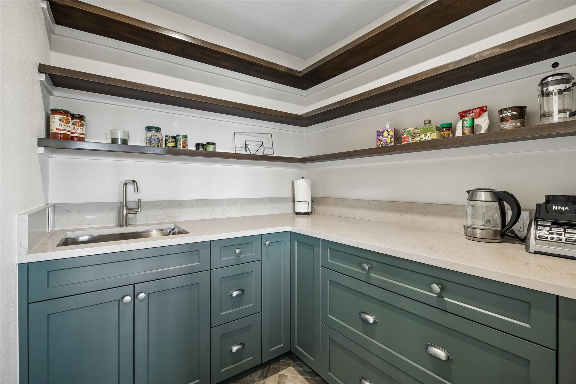 This kitchen area features striking dark green cabinetry paired with crisp white quartz countertops and a modern undermount sink. Above the counter, open wood shelving with matching trim adds warmth and texture against the neutral walls. The space is well-organized and functional, offering a clean, contemporary aesthetic with ample storage and prep space.