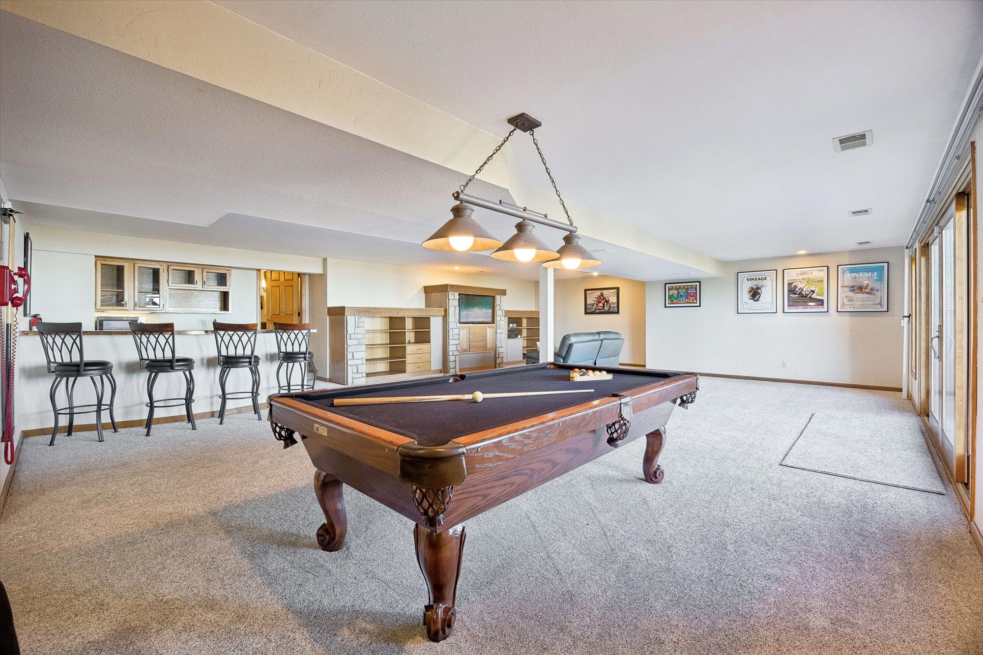 This spacious finished basement features a full-size pool table as its centerpiece, illuminated by a decorative hanging light fixture. The area also includes a custom wet bar with high-top seating and a cozy lounge space with integrated shelving and a television. The carpeted flooring and neutral color scheme create a comfortable and inviting atmosphere ideal for entertainment.