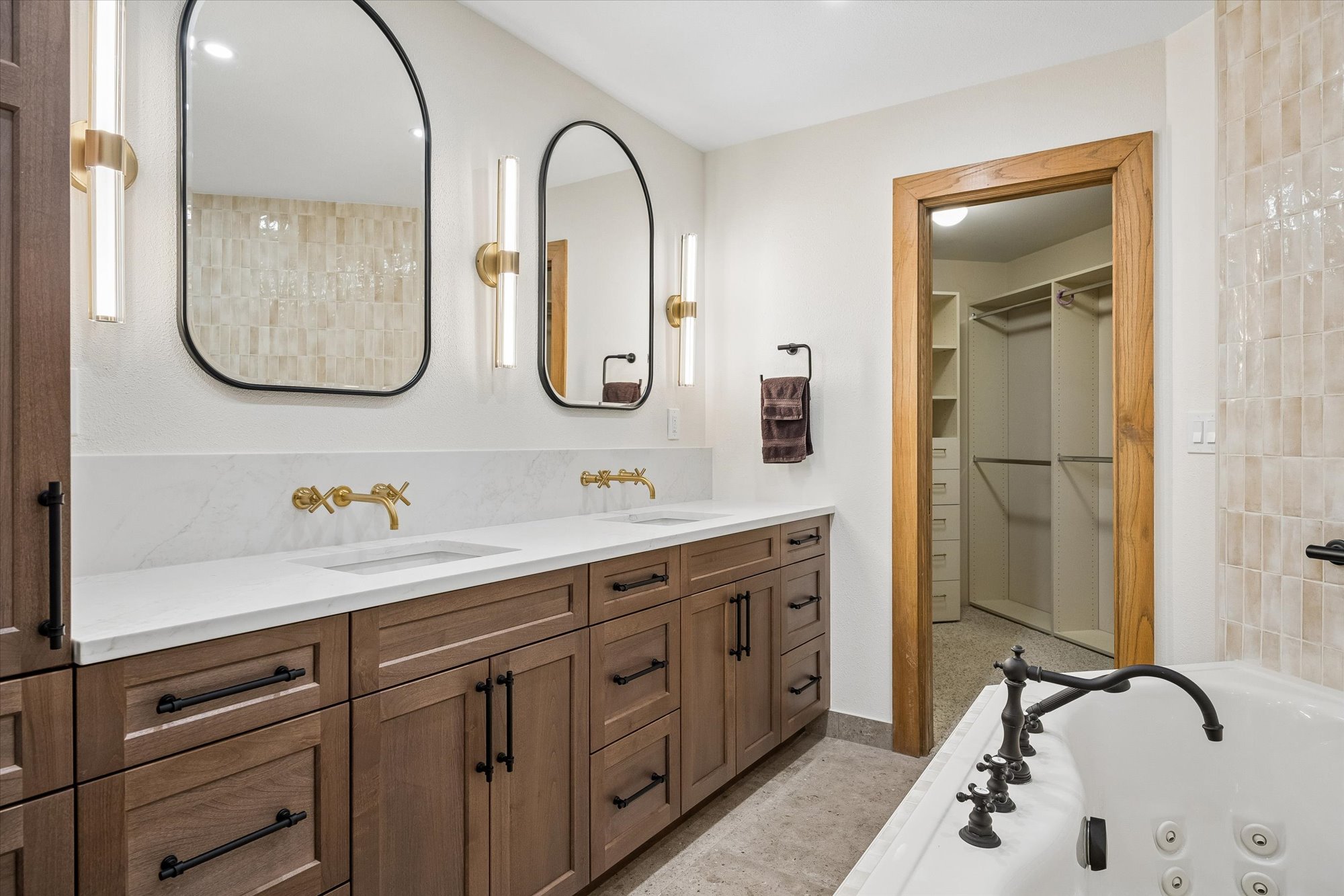 This primary bathroom features a sophisticated dual-vanity with rich wood cabinetry, white marble countertops, and stylish brushed gold fixtures. The space includes a jetted soaking tub and a direct view into a brightly lit, organized walk-in closet through a warm wooden doorway. The combination of modern wall-mounted lights and neutral tiled accents creates a luxurious and clean aesthetic.