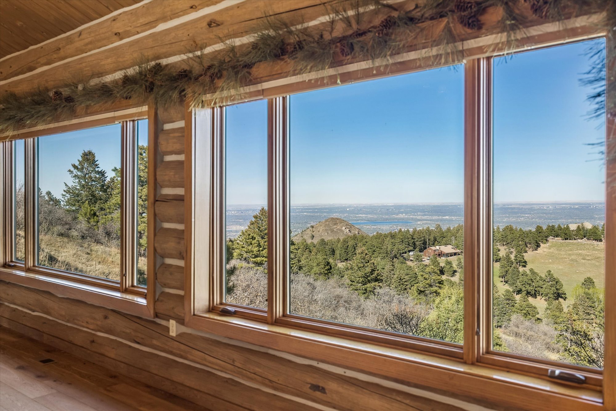 This interior view showcases the rustic charm of a log cabin, featuring detailed log walls and a natural wood aesthetic. Large, expansive windows provide panoramic, unobstructed views of the surrounding mountainous landscape and a distant valley, flooding the space with abundant natural light. The room is accented with tasteful holiday greenery, creating a warm, inviting atmosphere perfect for a mountain retreat.