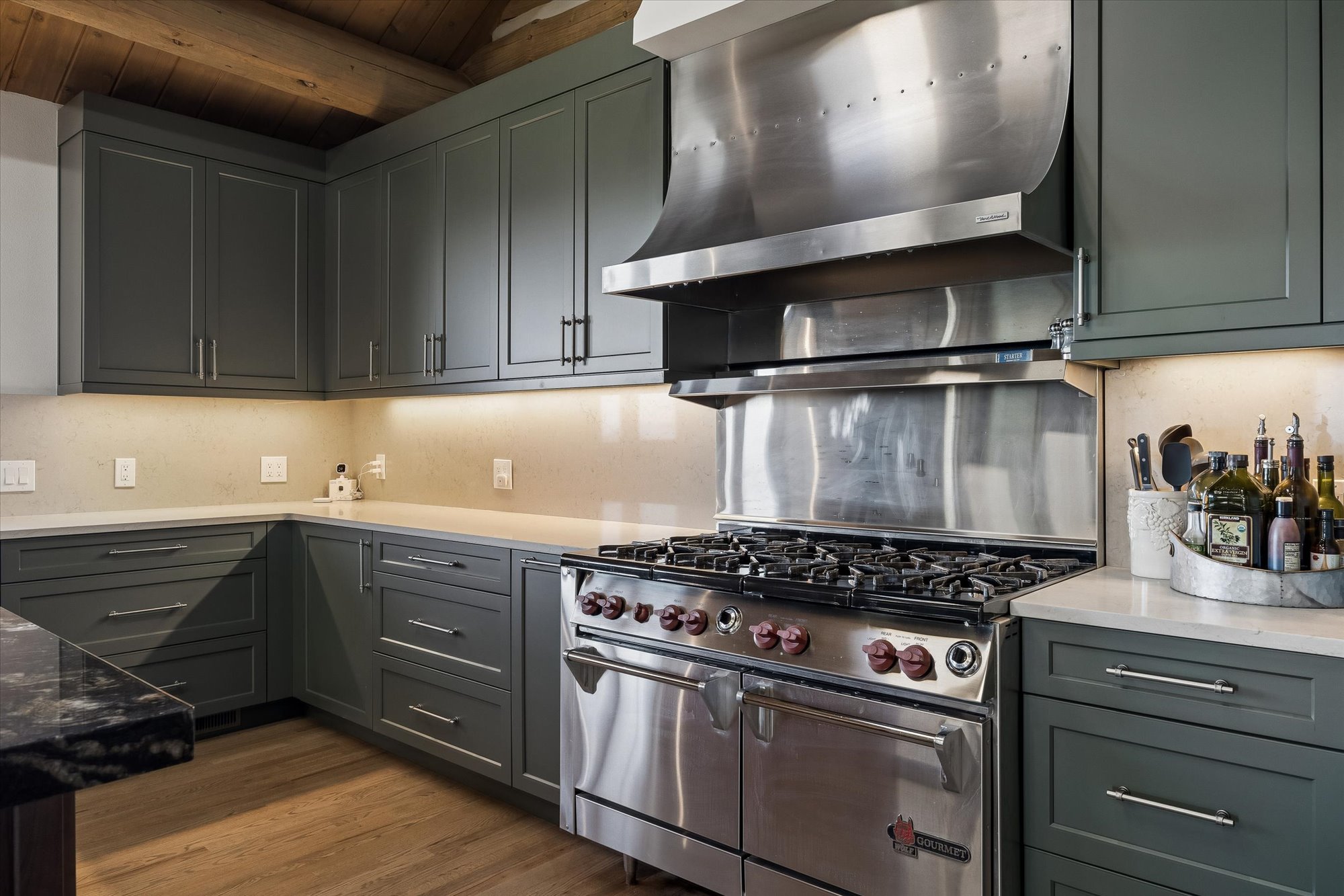This modern kitchen features elegant deep green cabinetry paired with sleek, professional-grade stainless steel appliances, including a high-end Wolf gas range. The space is well-coordinated with neutral quartz countertops, under-cabinet lighting, and clean hardware, creating a sophisticated and functional culinary area. The inclusion of light hardwood flooring and visible wooden ceiling beams adds a touch of warmth and texture to the contemporary design.