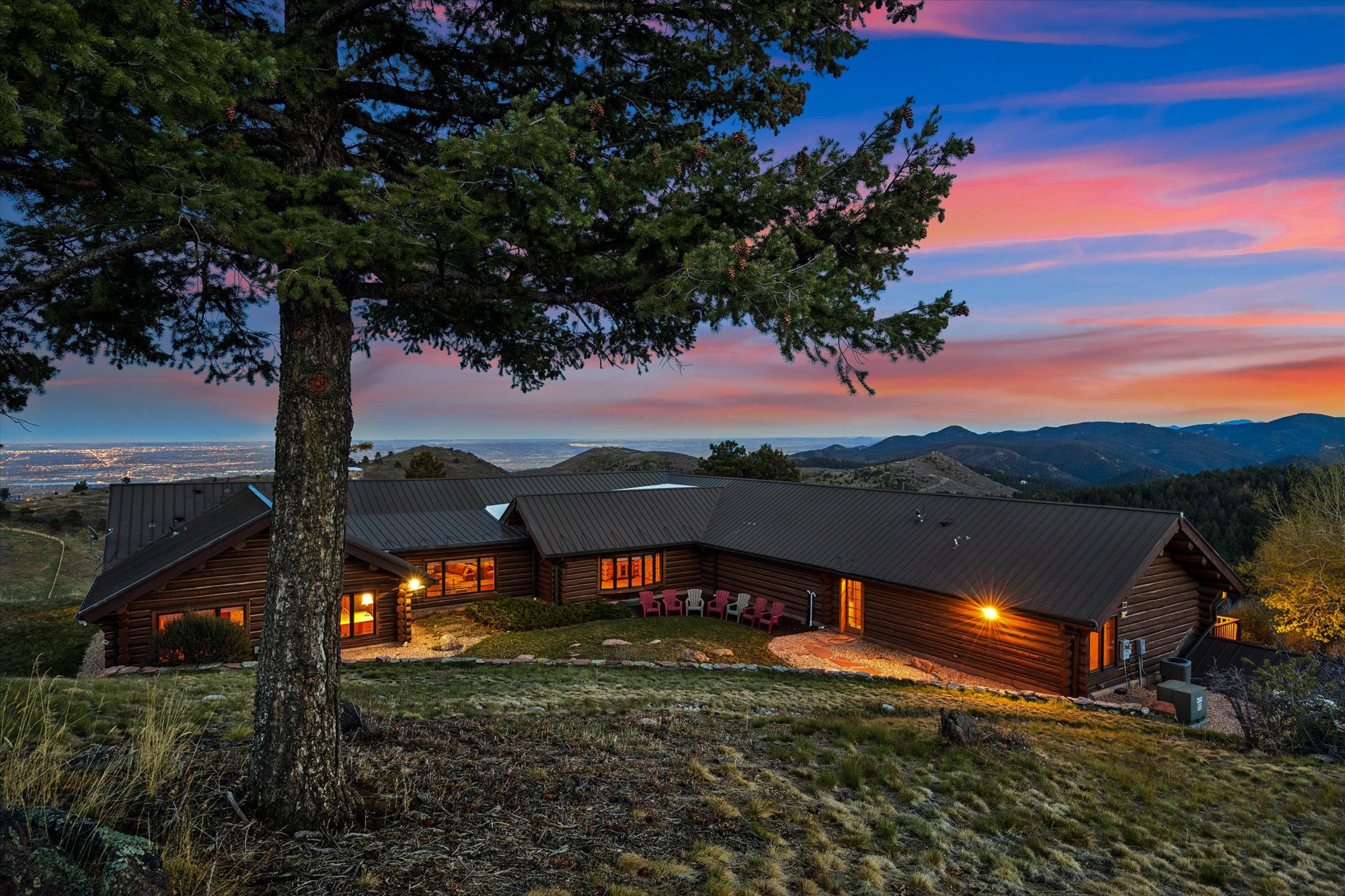 This stunning mountain cabin features a rustic log exterior with a durable metal roof, perfectly situated to overlook a panoramic landscape at twilight. The home includes a landscaped courtyard area with colorful patio seating, creating an inviting space to enjoy the breathtaking sunset and distant city lights. Its elevated, tiered design blends seamlessly with the natural terrain, highlighting the property's seclusion and scenic beauty.