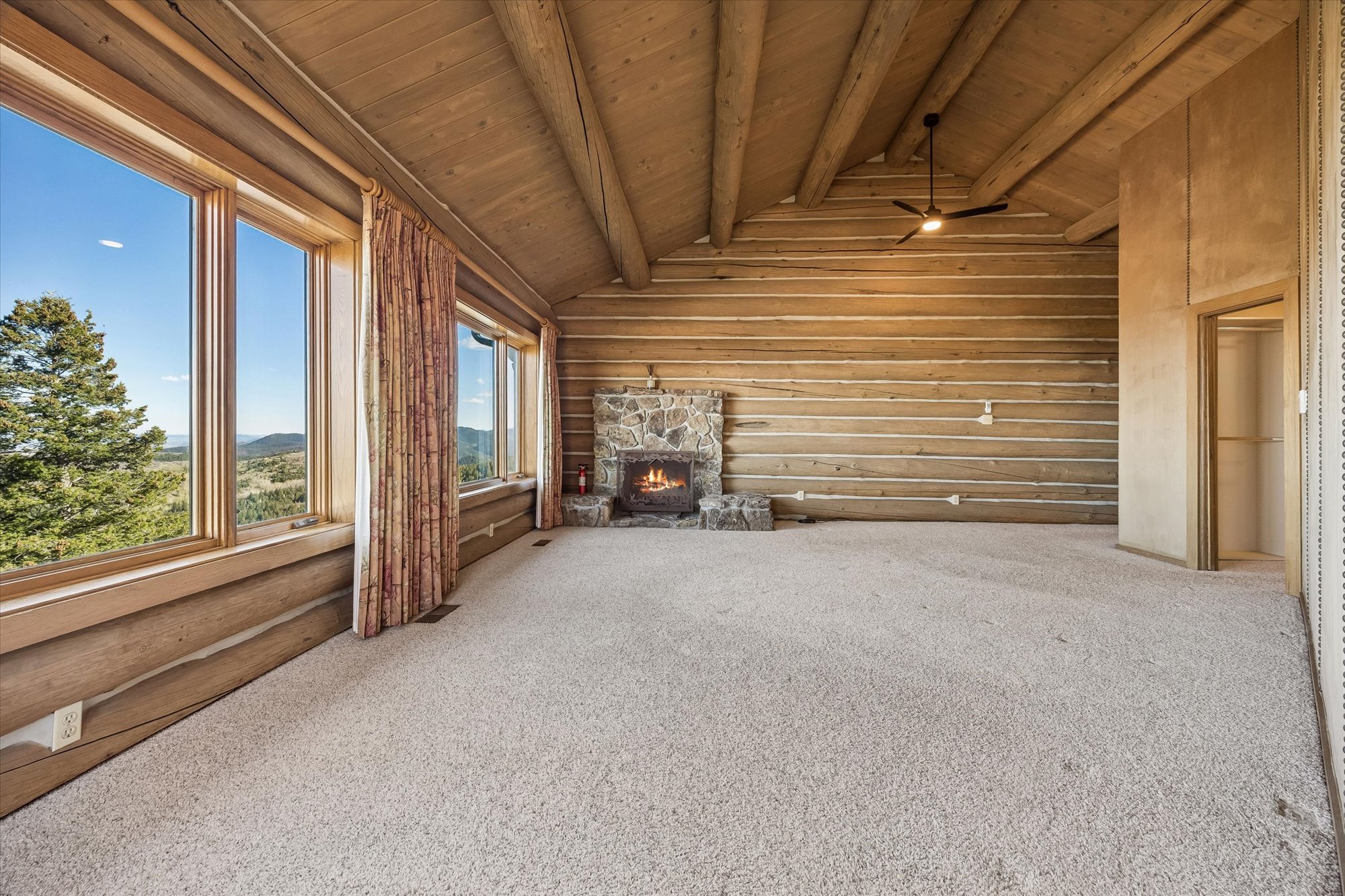 This spacious primary bedroom features rustic log cabin walls, exposed timber ceiling beams, and a cozy stone fireplace, creating a warm and inviting mountain lodge aesthetic. Large windows offer breathtaking panoramic views of the surrounding mountainous landscape, filling the room with natural light. The space is finished with neutral carpeting, providing a comfortable foundation for this elegant, high-altitude retreat.