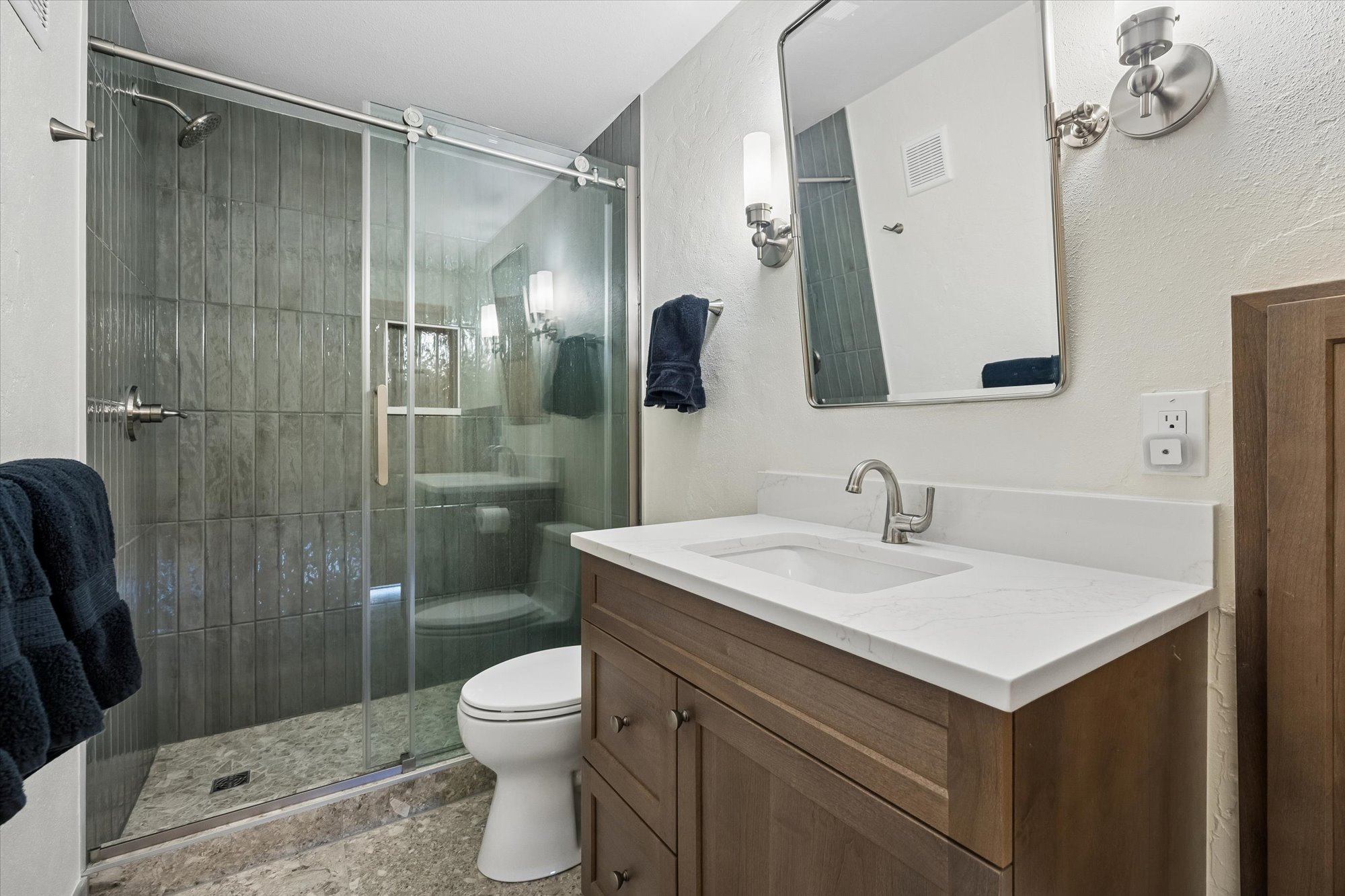 This contemporary bathroom features a wooden vanity with a white quartz countertop and a single faucet basin. A glass-enclosed shower showcases dark vertical tiling and a stone-look floor, adjacent to a white toilet. The space is brightened by a rectangular mirror with chic wall sconces and a minimalist aesthetic.