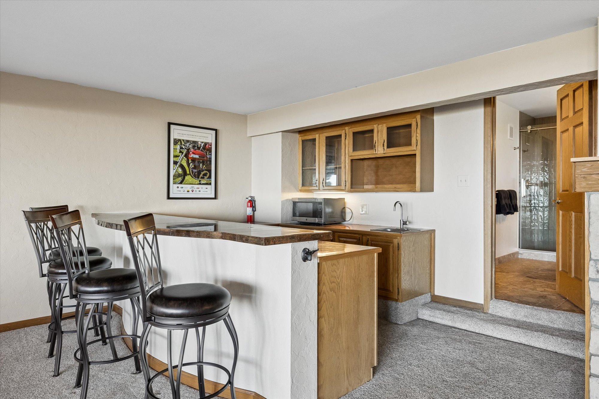 This interior basement area features a wet bar with wooden cabinetry, a built-in microwave, and a small sink, complemented by stone-topped bar counters and three matching stools. The space includes a designated wall for a framed decorative print, consistent beige wall paint, and access to an adjoining bathroom through a wooden door. The overall layout suggests a casual, traditional-style recreational space suitable for hosting guests.