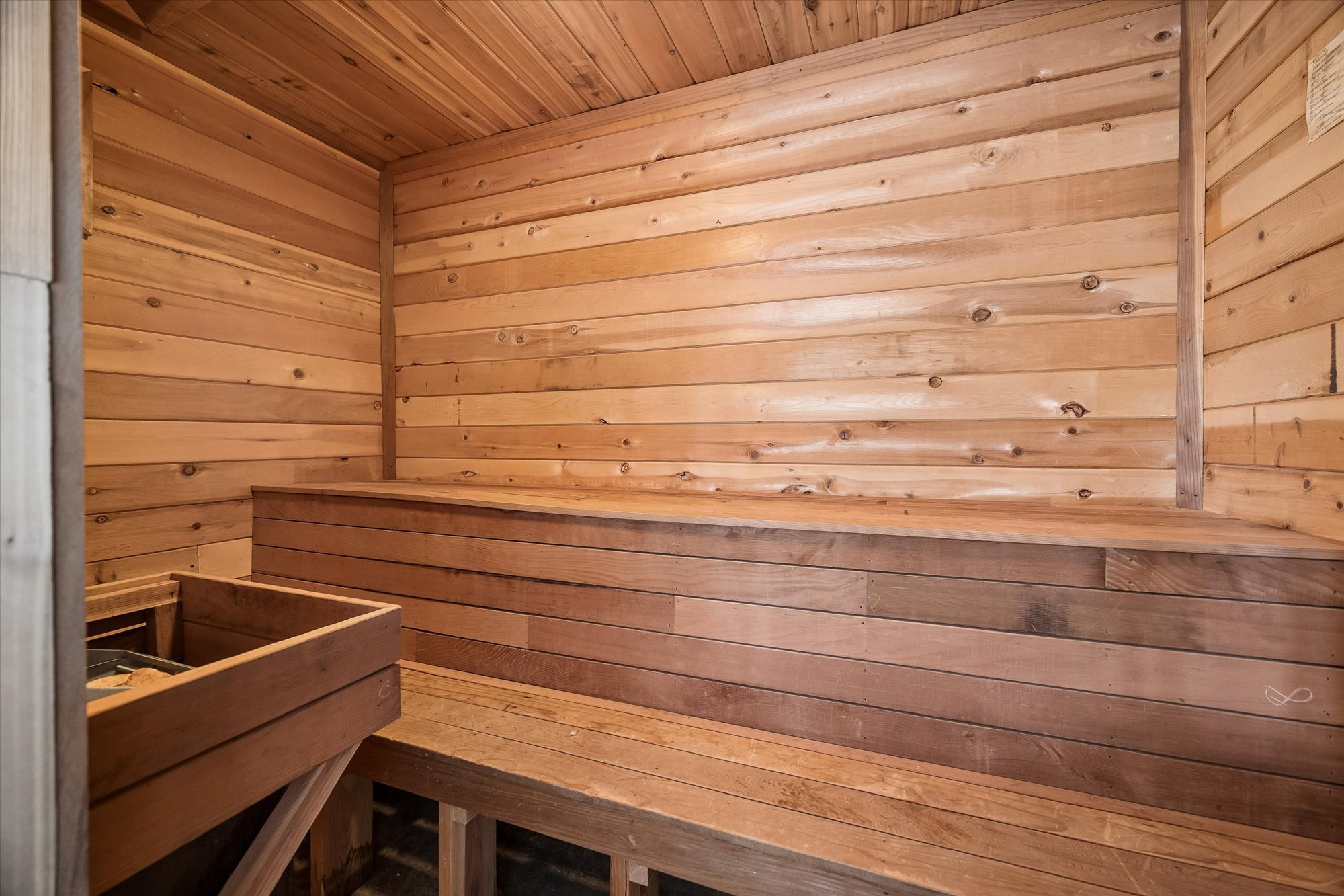 This image showcases a traditional sauna interior featuring warm-toned, natural wood paneling on the walls and bench seating. A wooden heater enclosure with stones is visible in the foreground, characteristic of a classic sauna setup. The wood-lined space provides a cozy, rustic, and spa-like atmosphere suitable for home wellness.