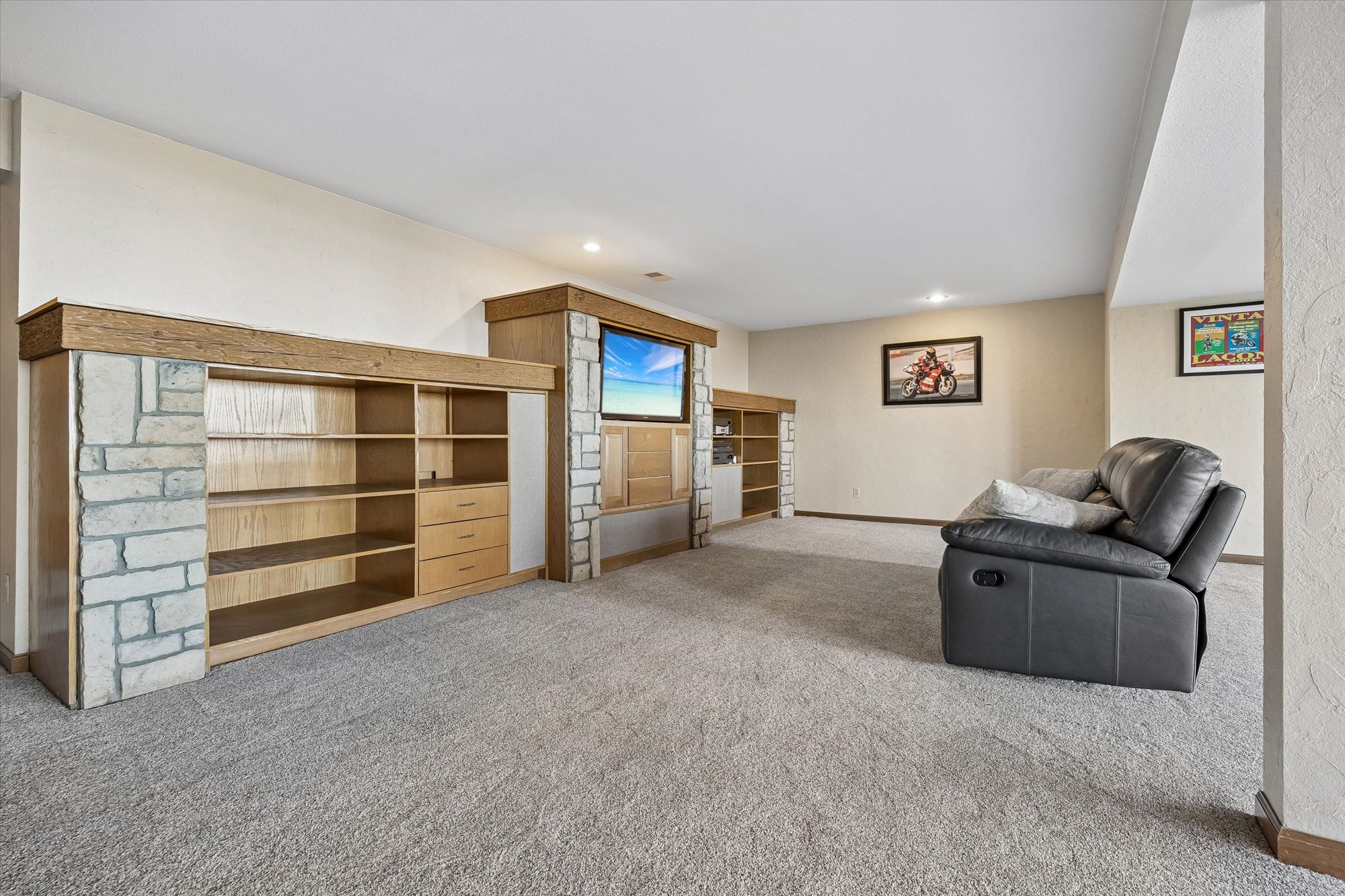 This finished basement area features a large built-in entertainment unit crafted with stone accents and wooden shelving. A plush gray recliner sofa provides comfortable seating facing the media center, while neutral carpet and wall tones create a warm, inviting atmosphere. The space is well-lit and decorated with framed wall art, making it an ideal family or recreational room.