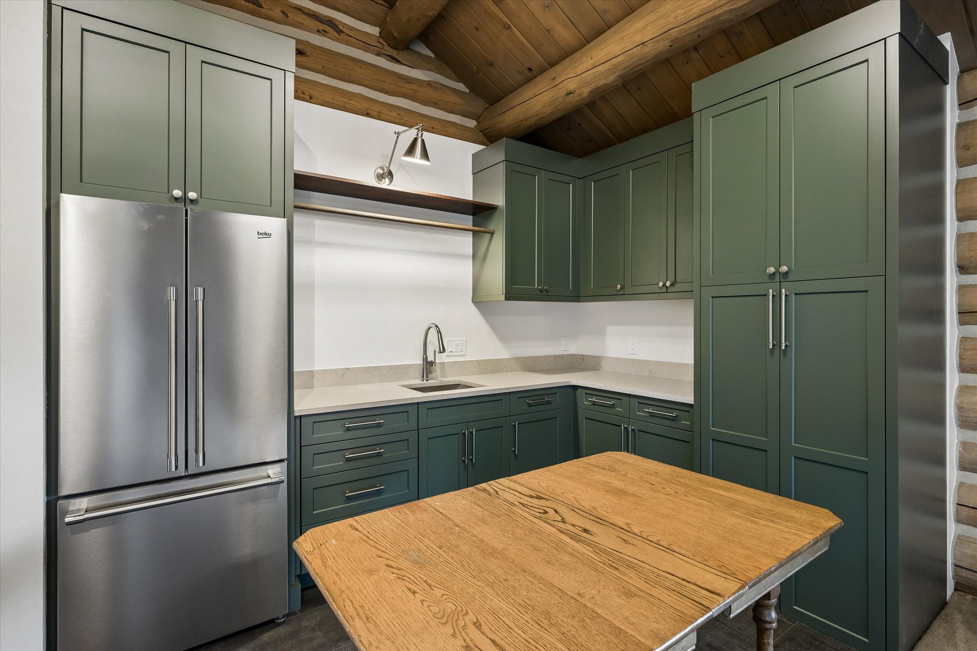 This kitchen features a distinct rustic-modern fusion with dark green cabinetry, stainless steel appliances, and sleek stone countertops. A unique architectural element is the exposed wooden beam ceiling, adding warmth to the space. The inclusion of a sturdy wooden table as a centerpiece and a minimalist shelf with integrated lighting provides a comfortable and functional aesthetic.