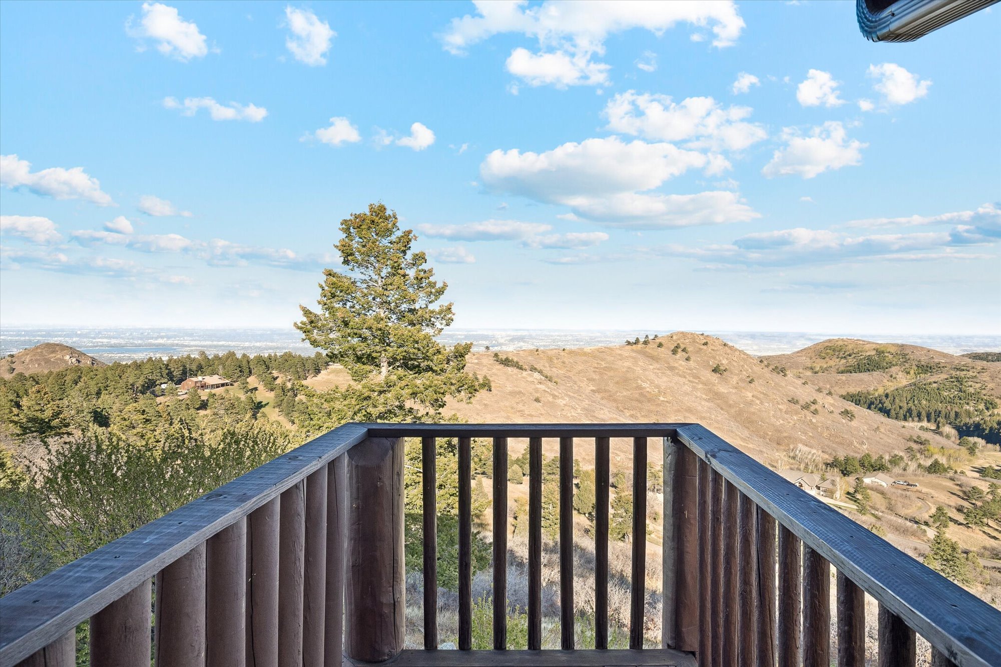 This image presents a deck with a rustic log-style railing that frames a wide, panoramic view of rolling hills and distant plains. The open design emphasizes the expansive, scenic outlook, making it an ideal space for outdoor relaxation or entertaining. The natural scenery and elevated perspective contribute to a serene and impressive atmosphere.