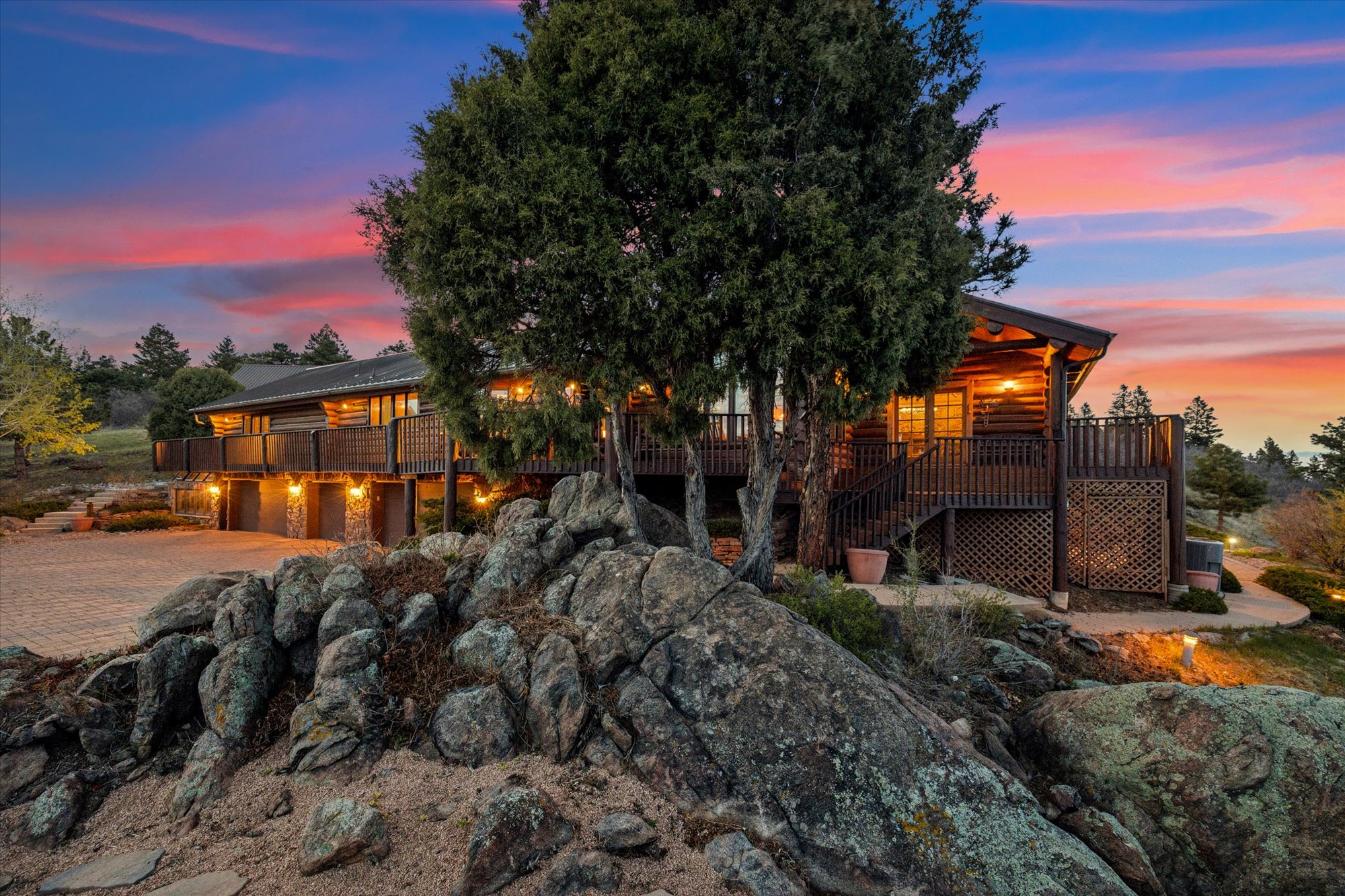 This is a beautiful log cabin-style home captured during a vibrant sunset, set against a backdrop of natural rock formations and greenery. The house features an expansive wooden deck that spans its length, above a multi-car garage entrance at the ground level. The warm glow of the interior lighting and exterior fixtures creates a welcoming and cozy atmosphere in this serene landscape.