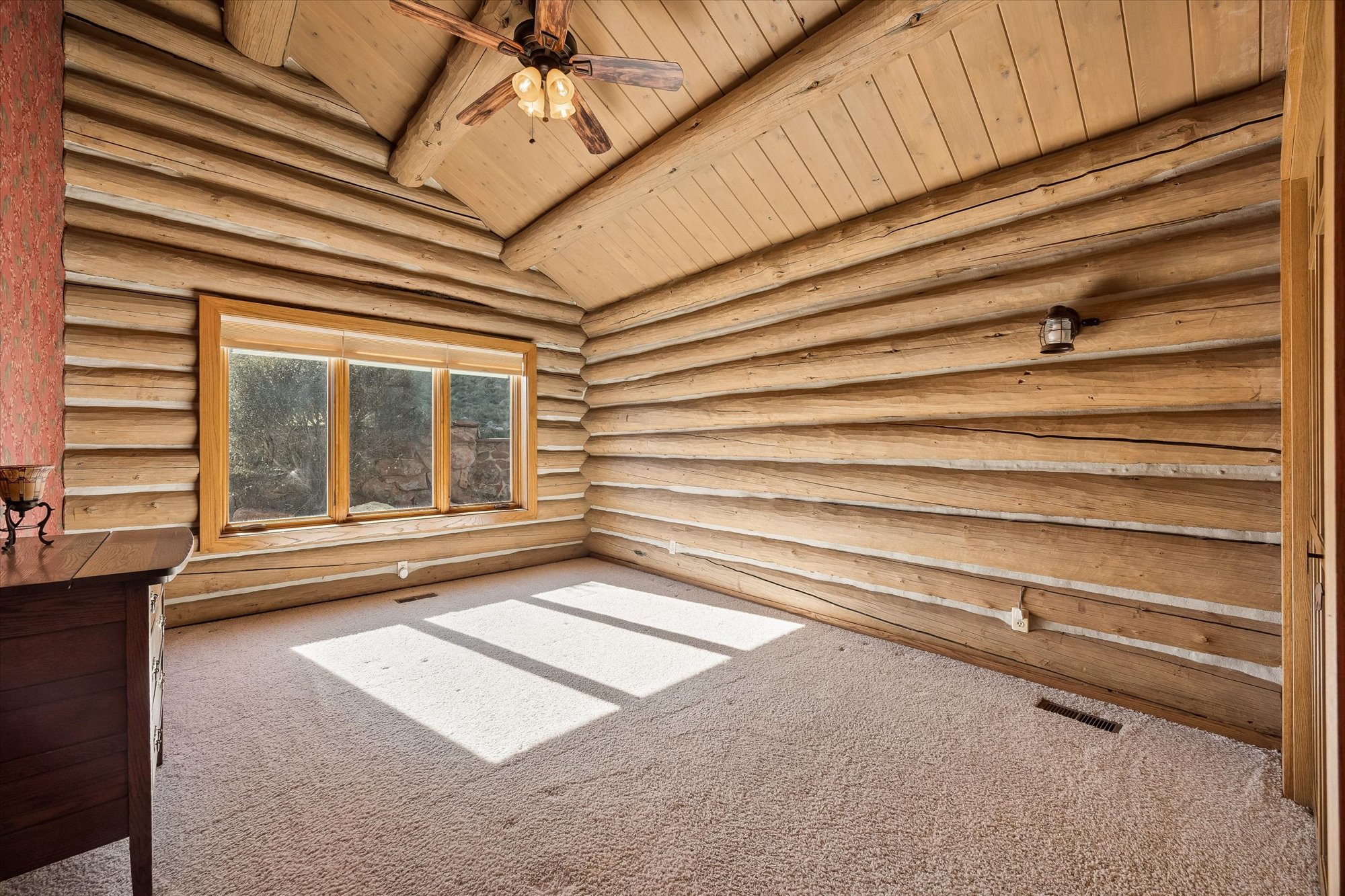 This rustic, cabin-style room features distinctive stacked log walls and a vaulted wooden ceiling, creating a warm, mountain-inspired aesthetic. The space is filled with natural light streaming through a large, triple-pane window and offers carpeting throughout. A wooden dresser and a vintage-style wall-mounted light fixture add to the cozy, traditional charm of this versatile living space.