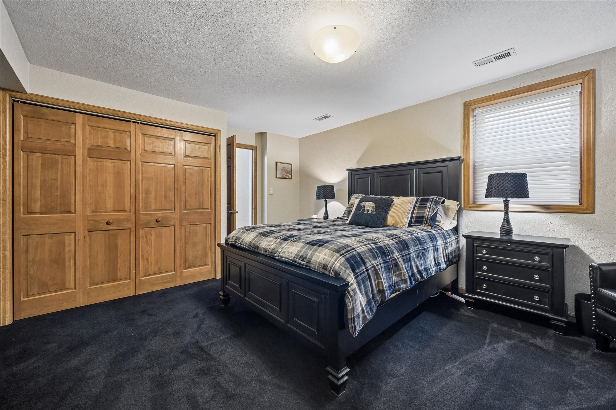 This guest bedroom features a dark-stained wood bed frame paired with matching nightstands and a neutral plaid bedding set. The room is grounded by dark carpeting and includes a spacious wooden closet with double doors, providing ample storage. The space is brightened by a large window with white blinds and a simple ceiling lighting fixture.