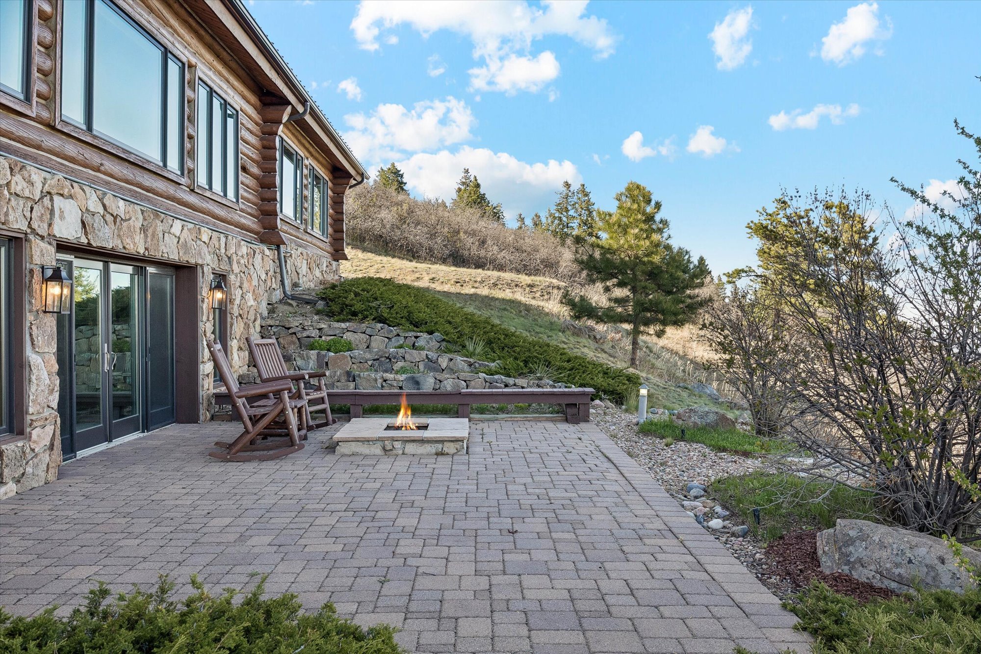 This outdoor patio space features a structured paver surface and a built-in stone fire pit, perfect for entertaining against a rustic mountain backdrop. A wooden bench and two matching rockers provide comfortable seating, while the side of the log home serves as an elegant, sturdy enclosure. The natural landscaping and sweeping scenic views create a tranquil and inviting atmosphere.
