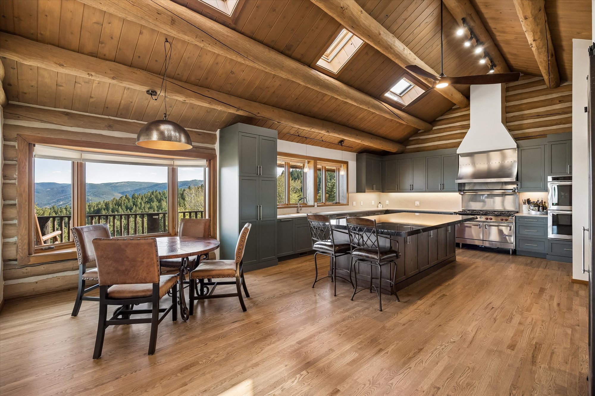 This rustic-style kitchen features vaulted wood ceilings with beams, skylights, and mountain views, blending warmth with modern amenities. It includes dark cabinetry, a large kitchen island with bar seating, and high-end stainless steel appliances, such as a professional-grade range and built-in wall oven. A cozy dining area with leather seating sits next to a large window, creating an inviting space perfect for both daily living and entertaining.