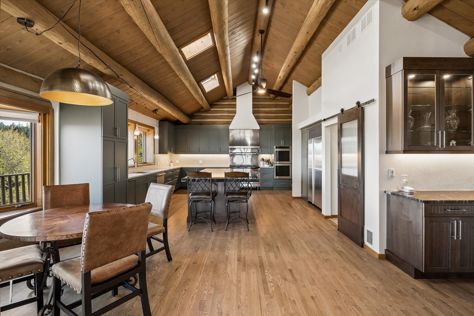 This spacious, open-concept kitchen seamlessly blends rustic charm with modern functionality, featuring exposed log ceiling beams and skylights. It is equipped with dark-toned cabinetry, high-end stainless steel appliances, and a central island with bar stool seating, while an adjacent dining area creates a warm, inviting space. The design is complemented by wood flooring and a sliding barn door, evoking a sophisticated mountain-lodge aesthetic.