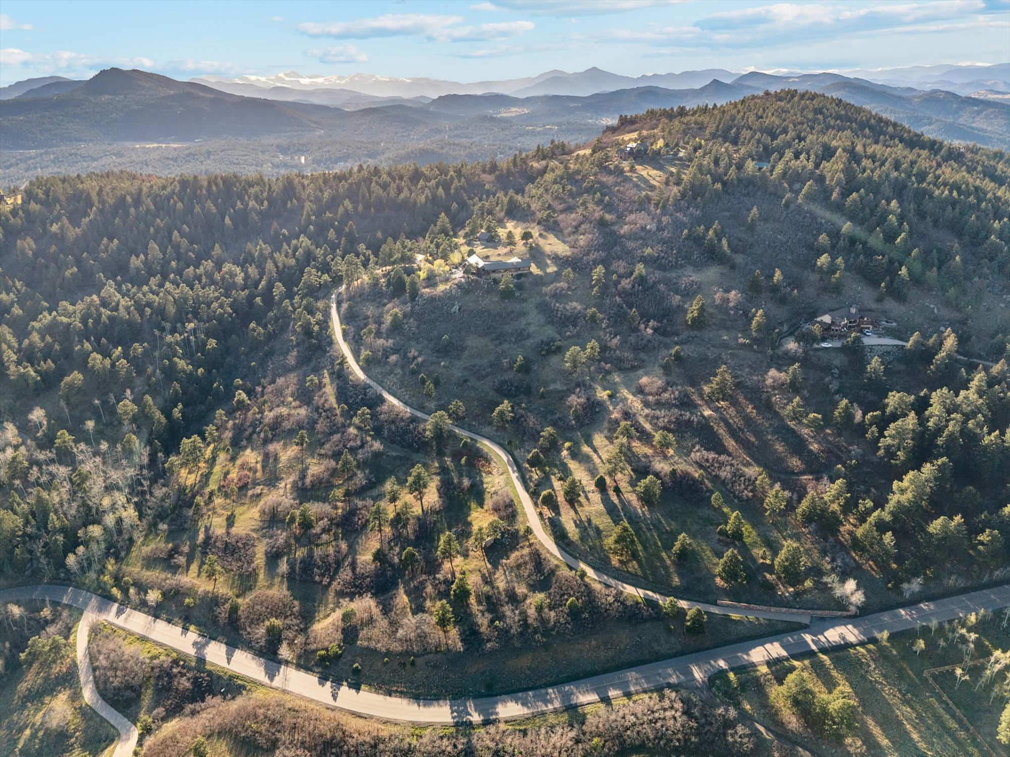 This stunning aerial view captures a hillside property set amidst a sprawling landscape of dense forest and mountainous terrain. A winding driveway gracefully curves through the wooded property, leading up to a large residence nestled near the crest of the hill. The scene highlights the tranquil seclusion, expansive natural beauty, and the picturesque setting of the home.