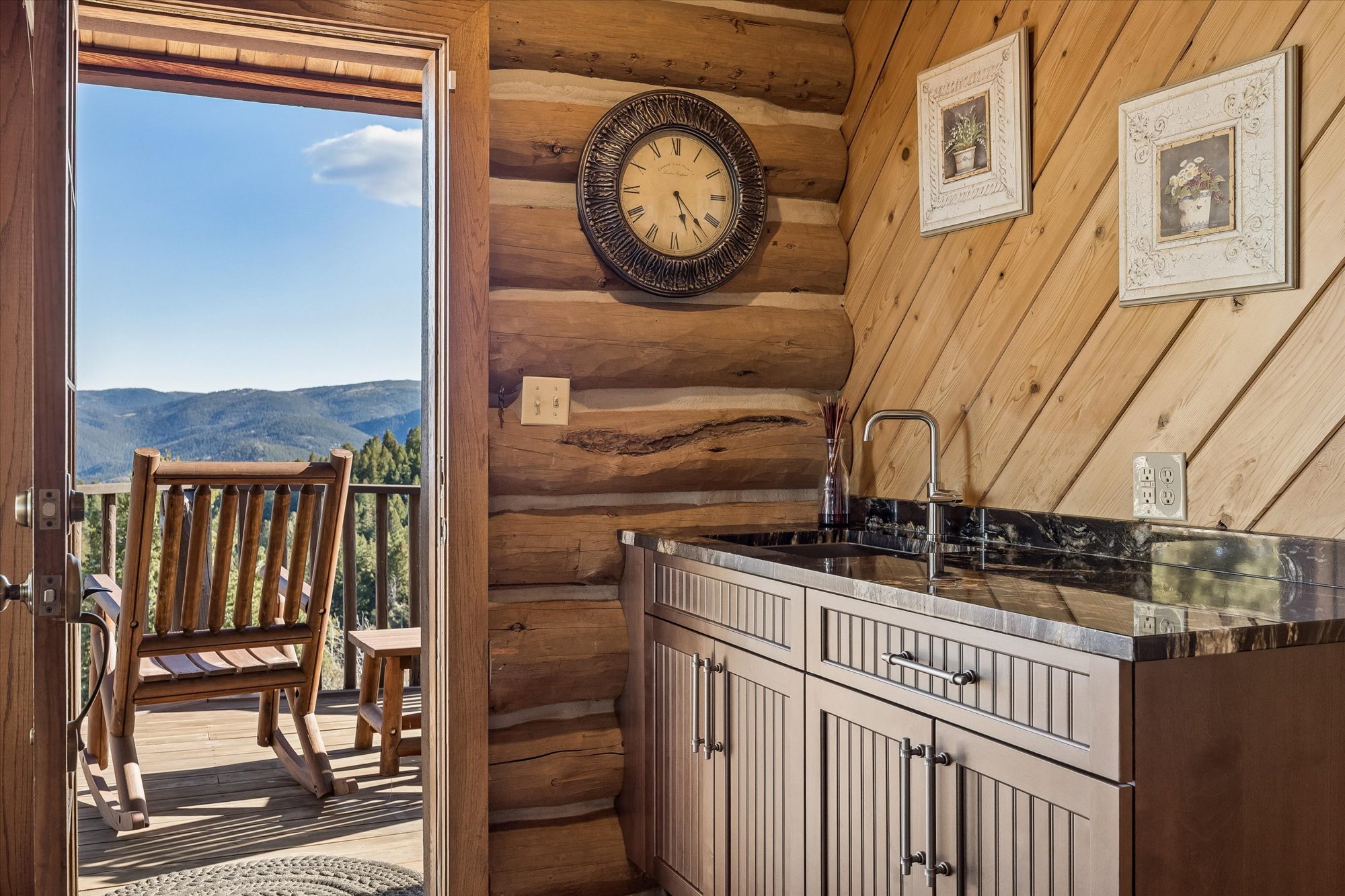 This interior image showcases a rustic wet bar area featuring warm, natural log walls and tongue-and-groove wooden paneling. The kitchenette includes a dark marble countertop with a stainless steel sink and modern cabinetry, while a door opens directly onto a deck with a scenic mountain view. A large antique-style wall clock and framed artwork add a refined, cozy touch to this charming woodland cabin space.