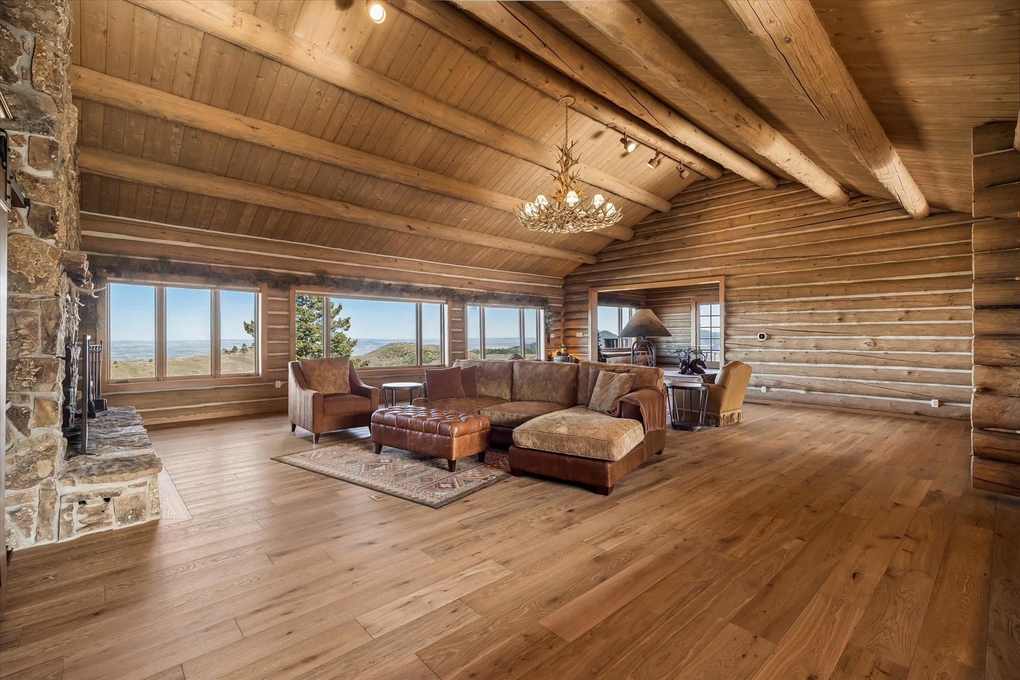 This spacious living room features authentic log walls, vaulted ceilings with exposed timber beams, and warm hardwood flooring, embodying a classic rustic mountain aesthetic. The room is centered around a large stone fireplace and a comfortable sectional sofa, all complemented by panoramic windows that frame stunning landscape views. An impressive antler chandelier serves as a focal point, enhancing the cabin's cozy and scenic atmosphere.