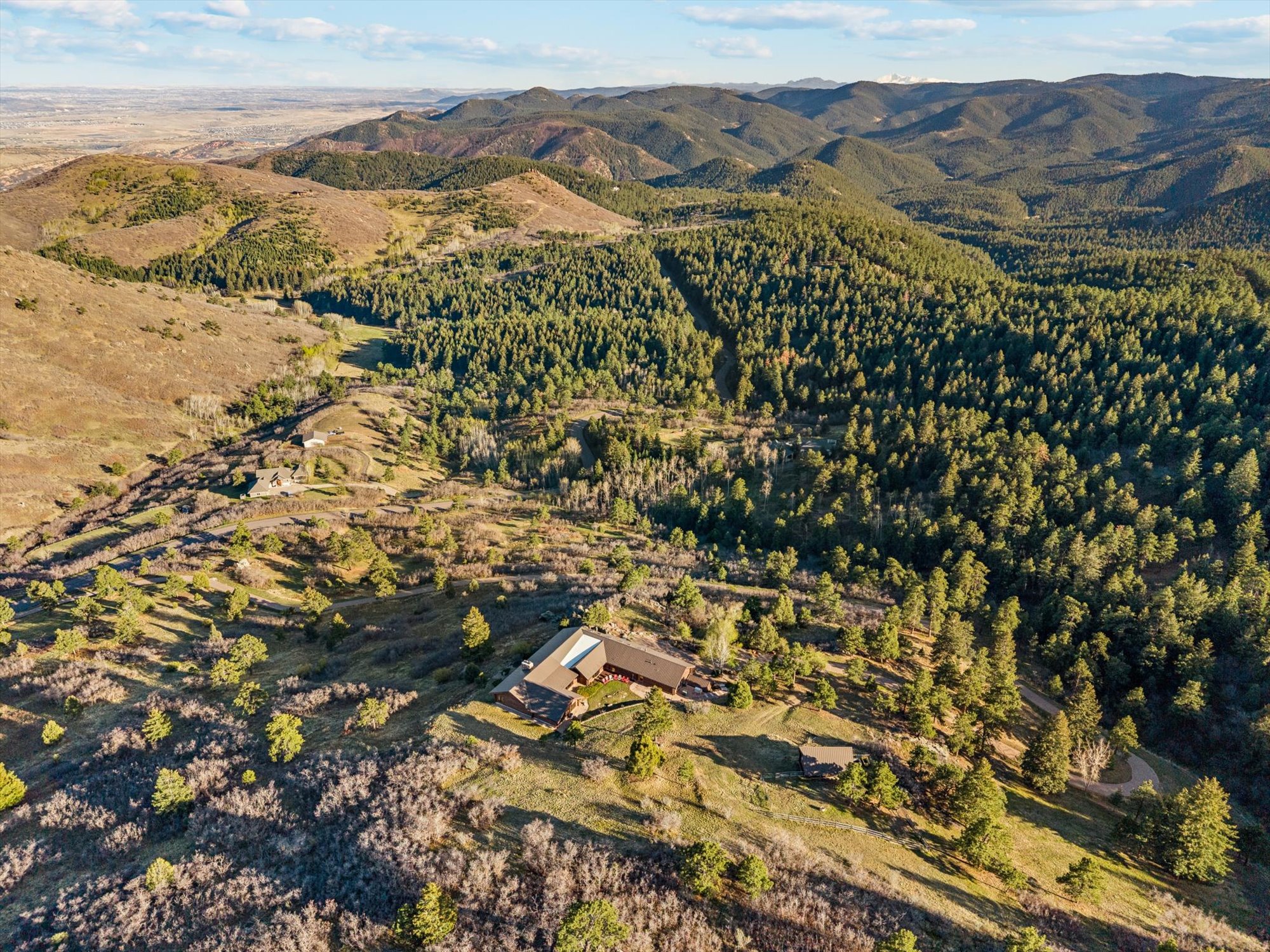 This panoramic aerial view showcases a sprawling residential property nestled within a mountainous, forested landscape. The main structure features a modern mountain-style design with a multi-level roofline, surrounded by expansive natural terrain, rolling hills, and dense evergreen forests. Its secluded setting offers significant privacy and a commanding perspective of the surrounding wilderness.