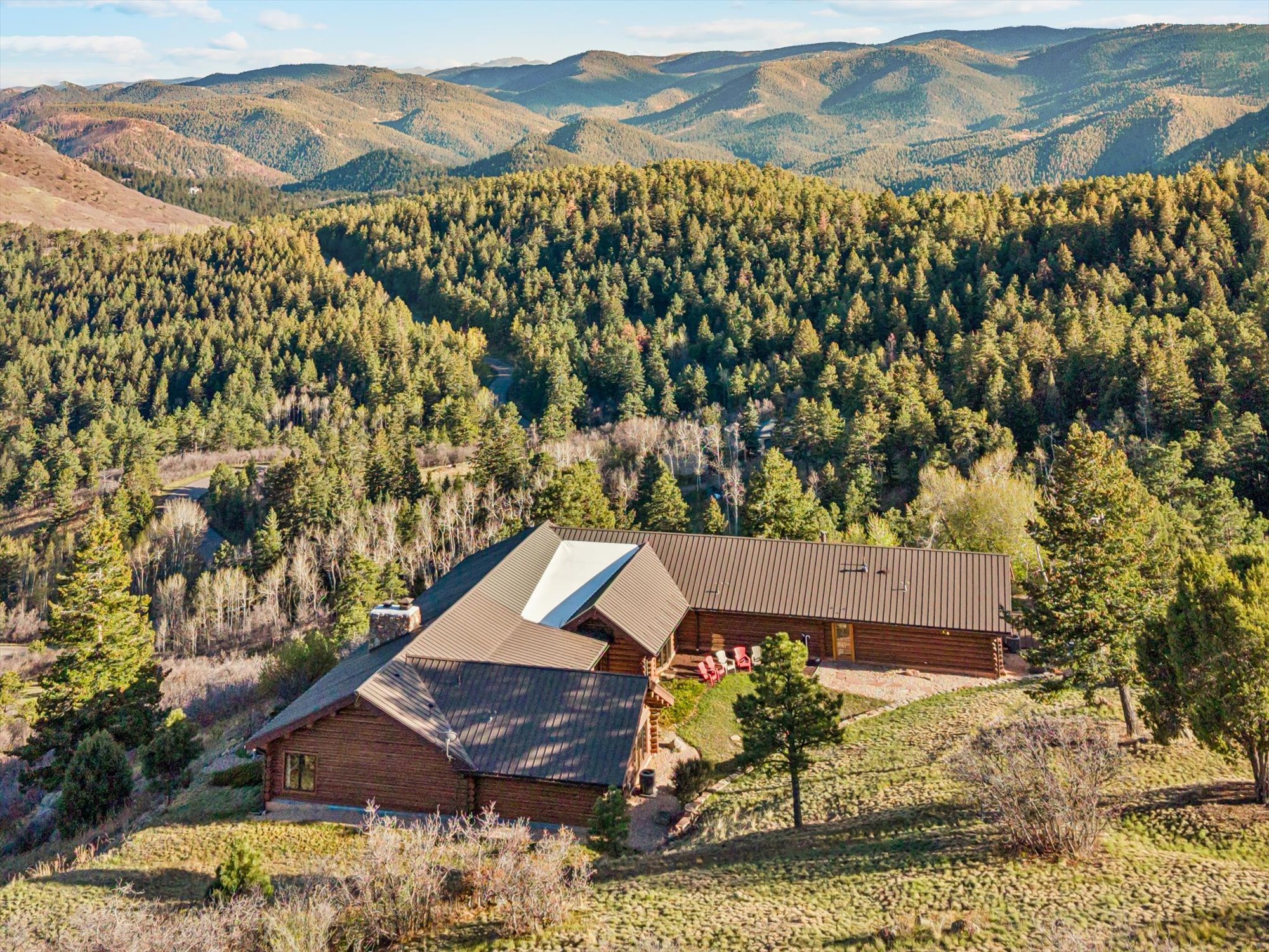 This aerial view showcases a rustic log cabin nestled in a vast, mountainous forested landscape. The home features a complex metal roofline with varied angles, a stone chimney, and a cozy patio area with outdoor seating. The setting offers a serene, secluded atmosphere with expansive panoramic views of the surrounding hills and dense evergreen forest.