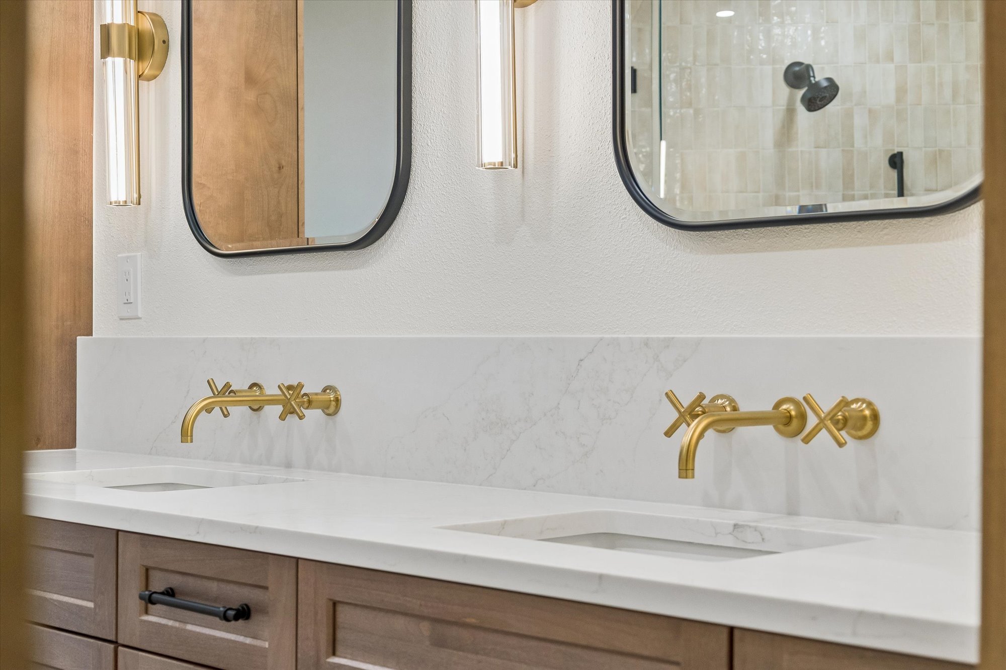 This modern primary bathroom features a sleek double vanity with custom wood cabinetry and a clean white marble countertop. Highlights include brushed gold wall-mounted faucets, stylish arched mirrors, and elegant linear sconce lighting that create a sophisticated, high-end feel.