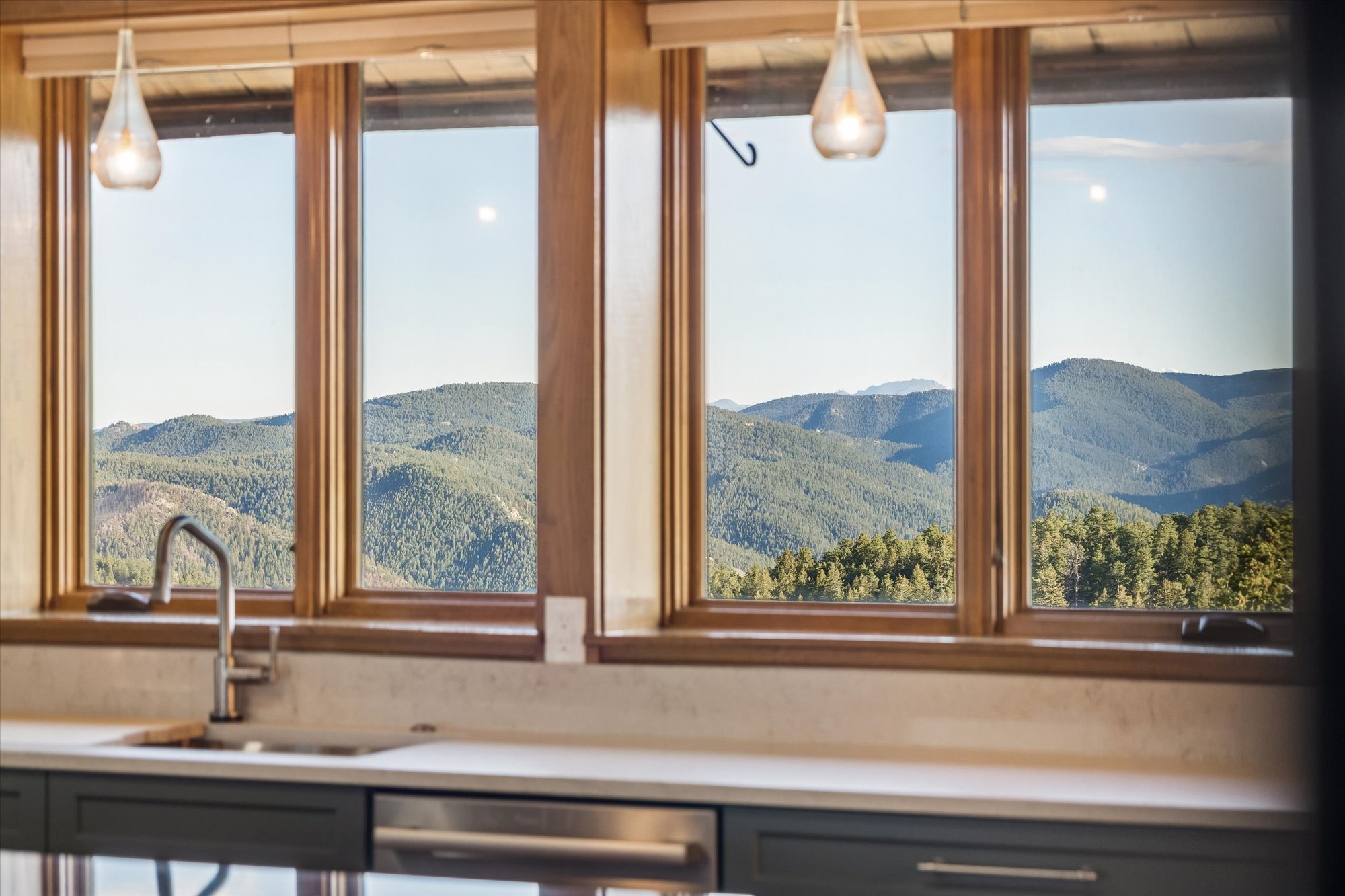 This interior kitchen view highlights a sleek, modern workstation featuring dark cabinetry, a light-colored countertop, and a stainless steel faucet. The standout feature is the large, multi-pane picture window that frames a breathtaking, expansive view of forested mountains. Elegant glass pendant lights hang above, enhancing the inviting atmosphere of this scenic space.