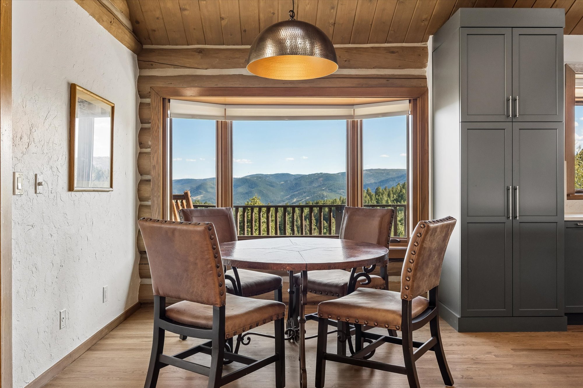 This cozy dining area features a round copper-top table paired with four leather chairs with nailhead trim, creating a warm, mountain-rustic aesthetic. A stylish industrial-style dome pendant light hangs above the set, while a large bay window offers breathtaking panoramic views of the forested mountains. The space is anchored by natural log walls on one side and a sleek, tall gray storage cabinet on the other, blending modern design with a classic lodge atmosphere.
