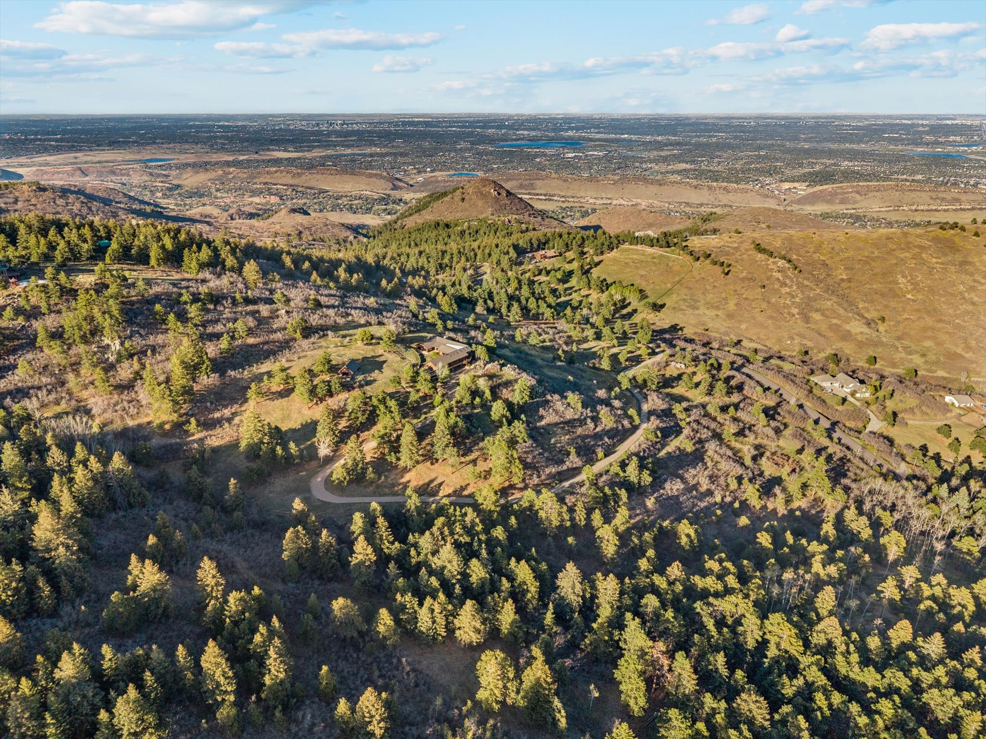 This aerial view showcases a hillside property nestled within a wooded, mountainous landscape. A private gravel driveway winds through the trees toward a large residential estate, while the background reveals a vast, expansive city view stretching toward the horizon. The setting offers a sense of privacy and seclusion while maintaining proximity to the urban area.