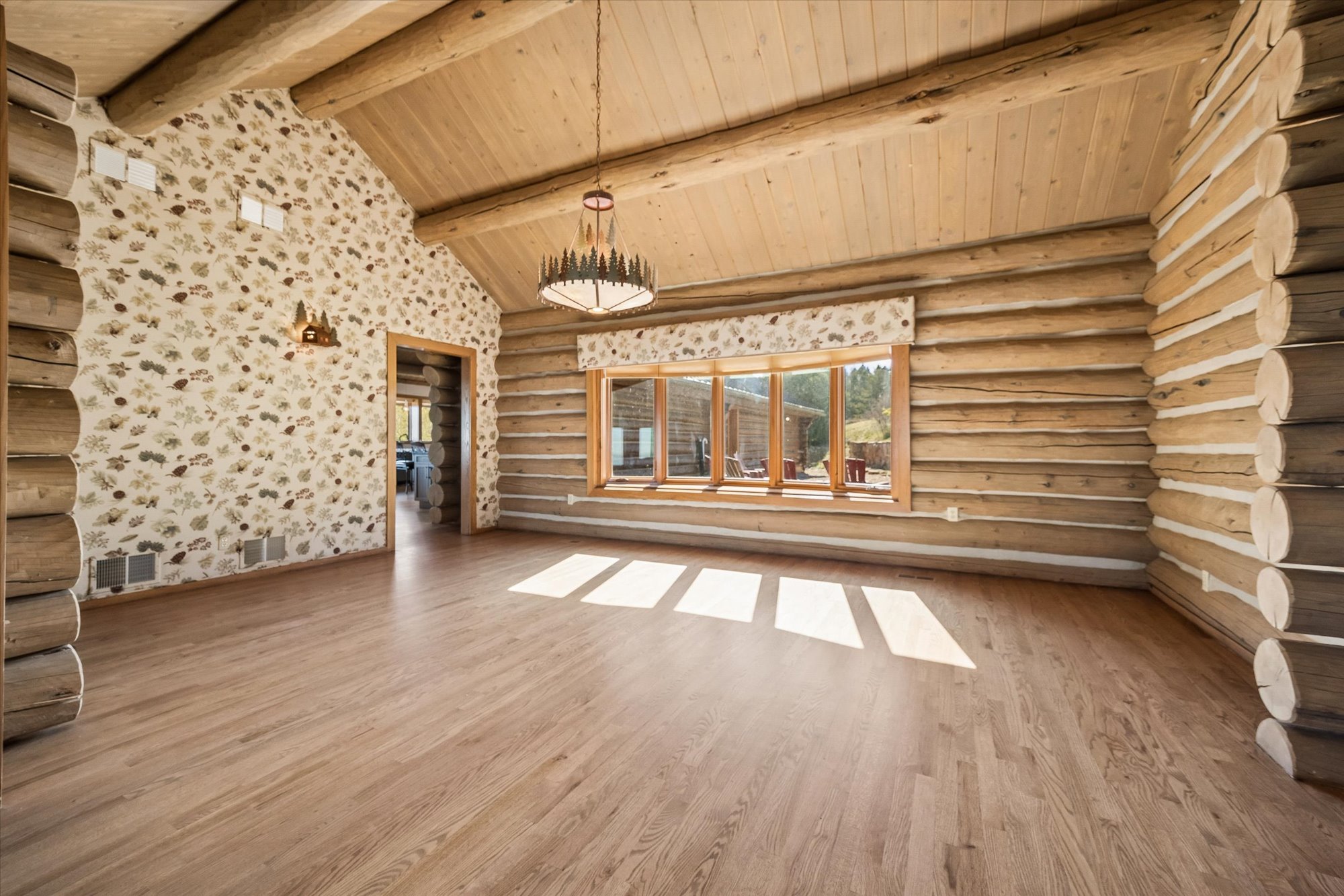 This spacious, open-concept living area features classic log cabin construction with exposed timber beams and horizontal log walls. The room is accented by a decorative, leaf-patterned wallpaper on one wall and a rustic chandelier above, with large windows flooding the space with natural light. It offers a warm, inviting atmosphere characteristic of a cozy mountain retreat.