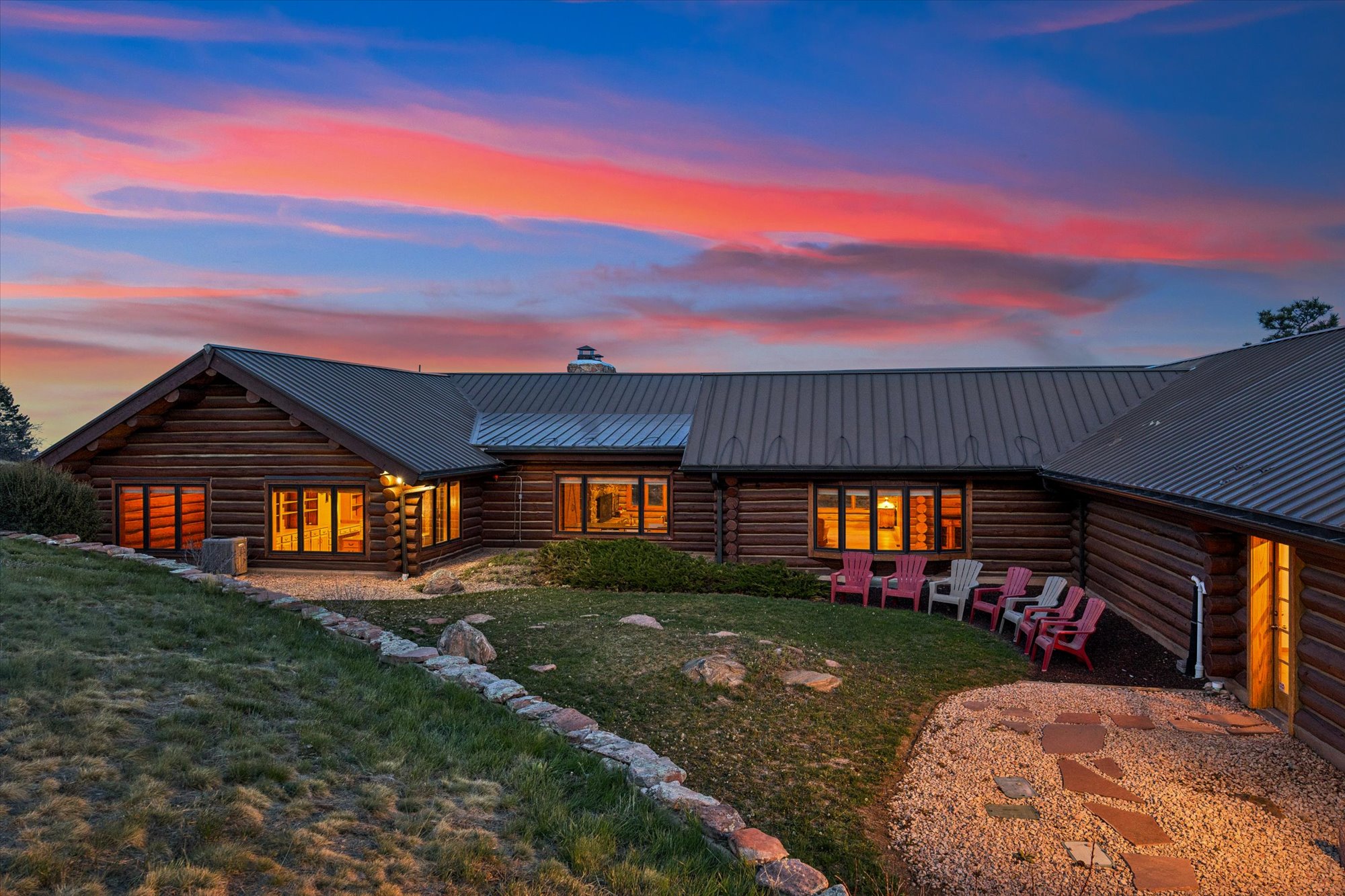 This charming, sprawling log cabin home is showcased at sunset, featuring a classic rustic design with a metal roof and glowing interior windows. A well-maintained courtyard lawn sits in the center of the structure, bordered by a stone retaining wall and an inviting gravel sitting area arranged withadirondack chairs. The warm lighting against the dramatic, colorful sky creates a welcoming and picturesque outdoor living space.