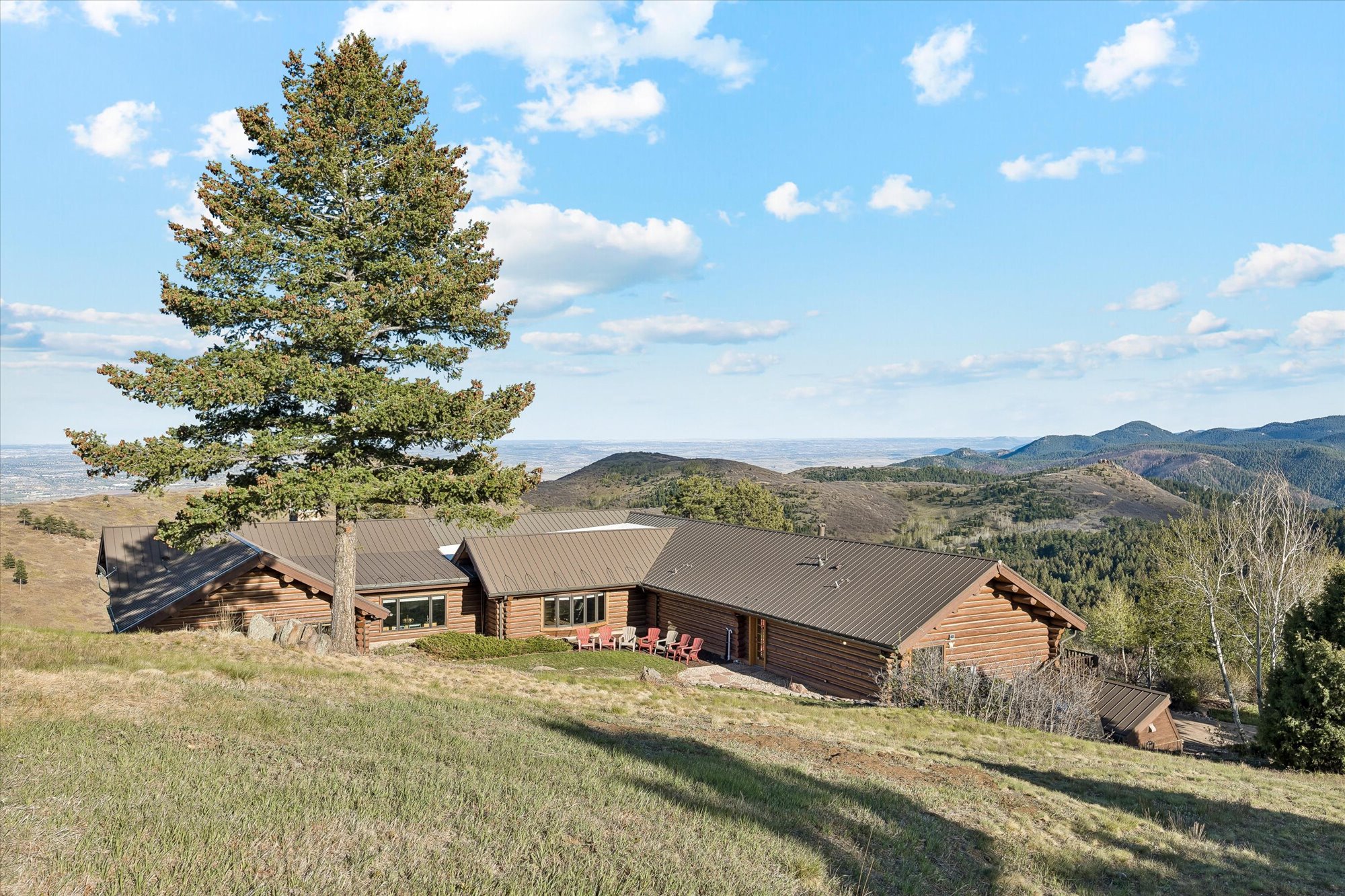 This scenic mountain residence features a rustic log-cabin exterior design with a modern, low-profile metal roof. The property is situated on a hillside, offering breathtaking panoramic views of the surrounding valley and distant mountain ranges. A small patio area sits outside, and a large, mature pine tree anchors the front landscape, enhancing the serene and natural setting.